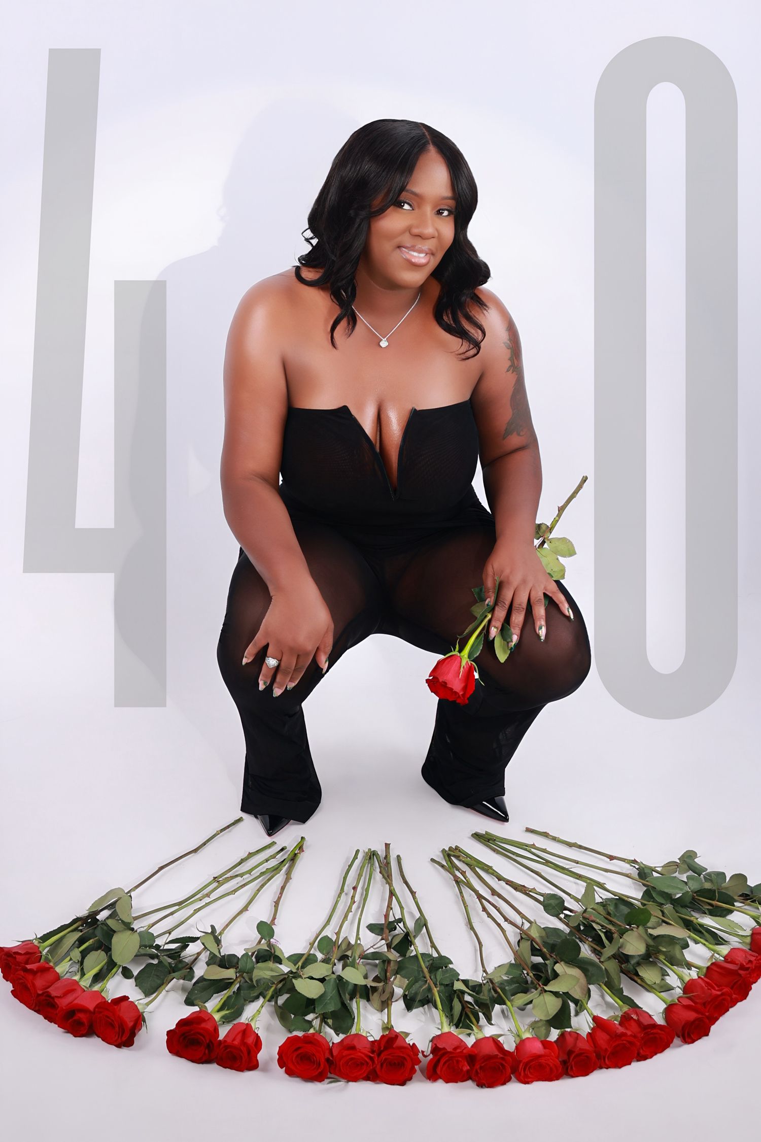 Confident Black woman in black outfit crouches with 40 red roses for birthday celebration.