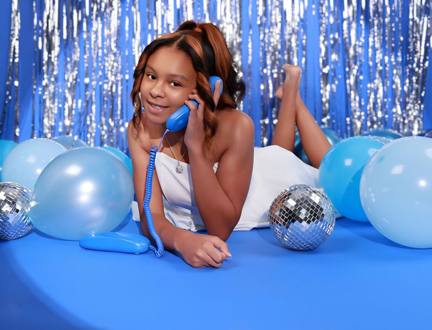 Young girl in white dress posing with blue balloons and disco balls on blue backdrop.
