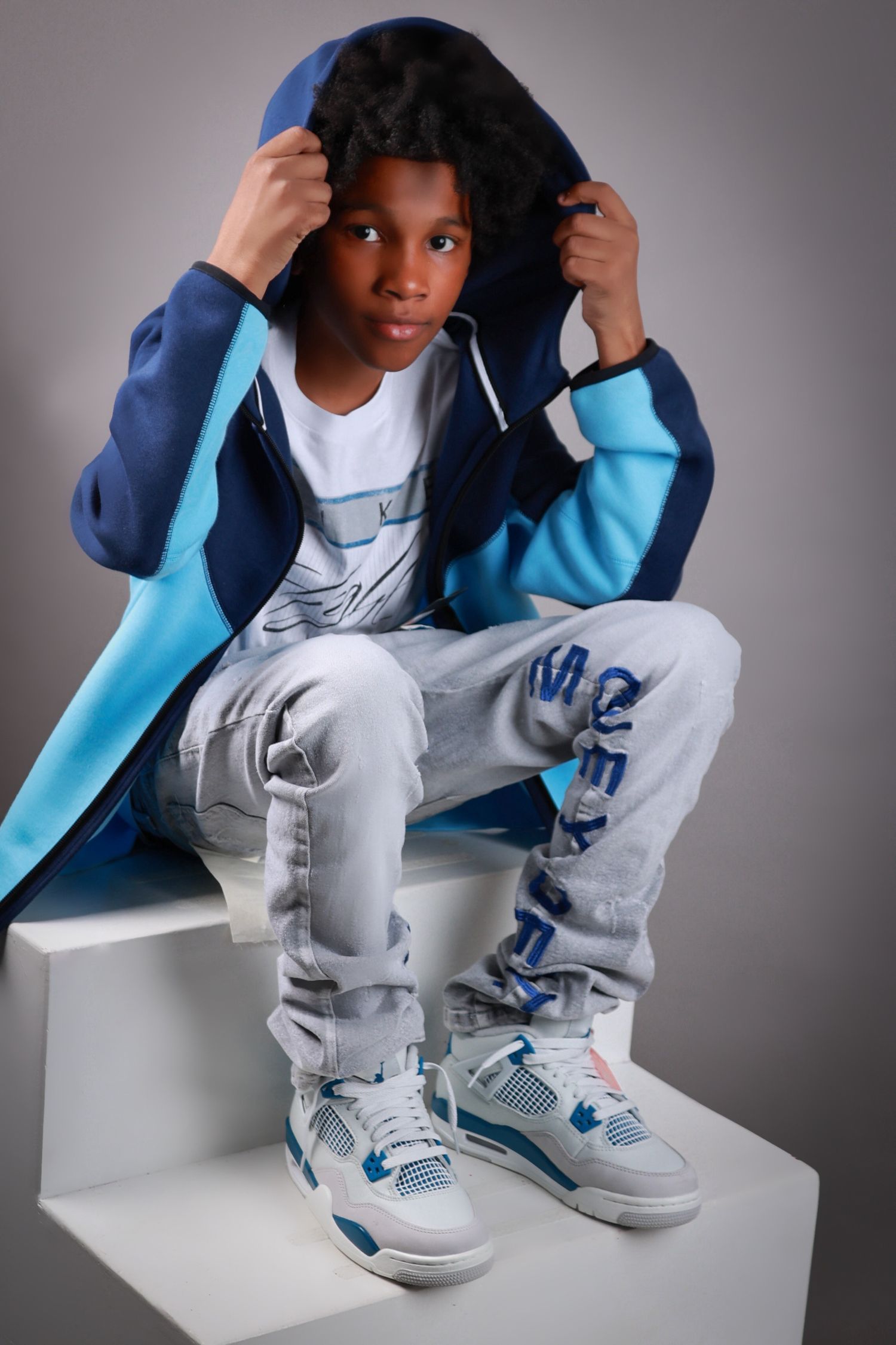Young boy in blue hoodie and Jordan sneakers poses on white steps in studio.
