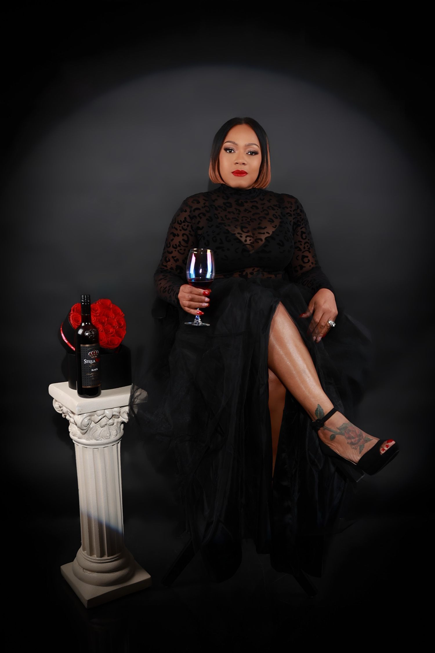 Woman in elegant black lace gown seated beside a pedestal with wine and red roses.