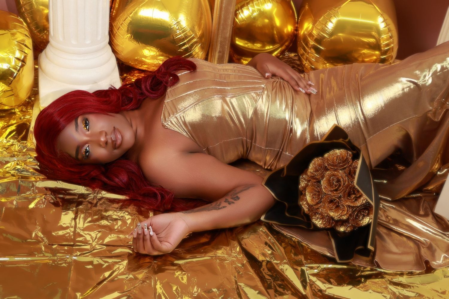 Woman in gold dress with red hair poses among gold balloons and metallic fabric.