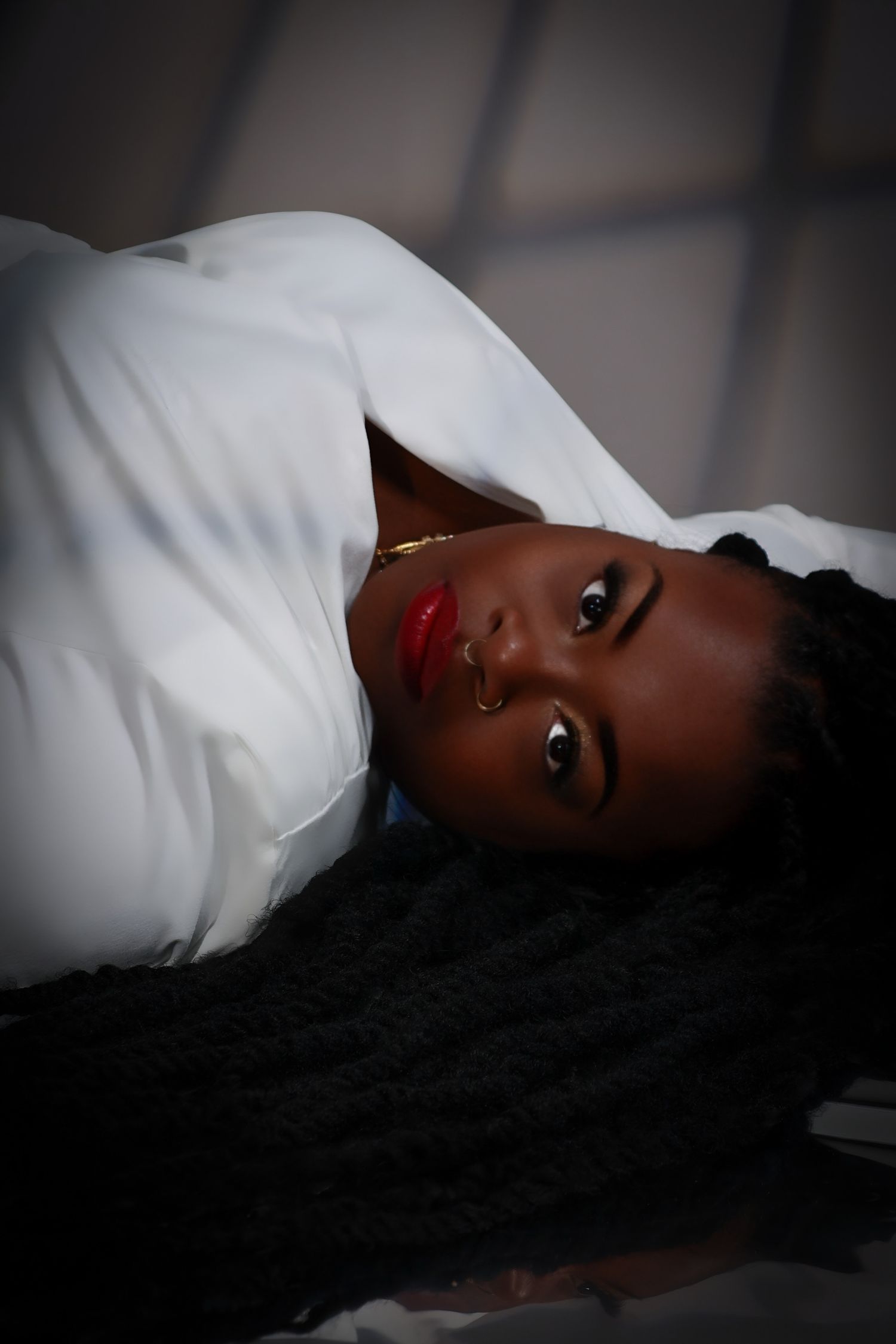 Young woman in white top lying down with red lips and braided hair in dramatic lighting.