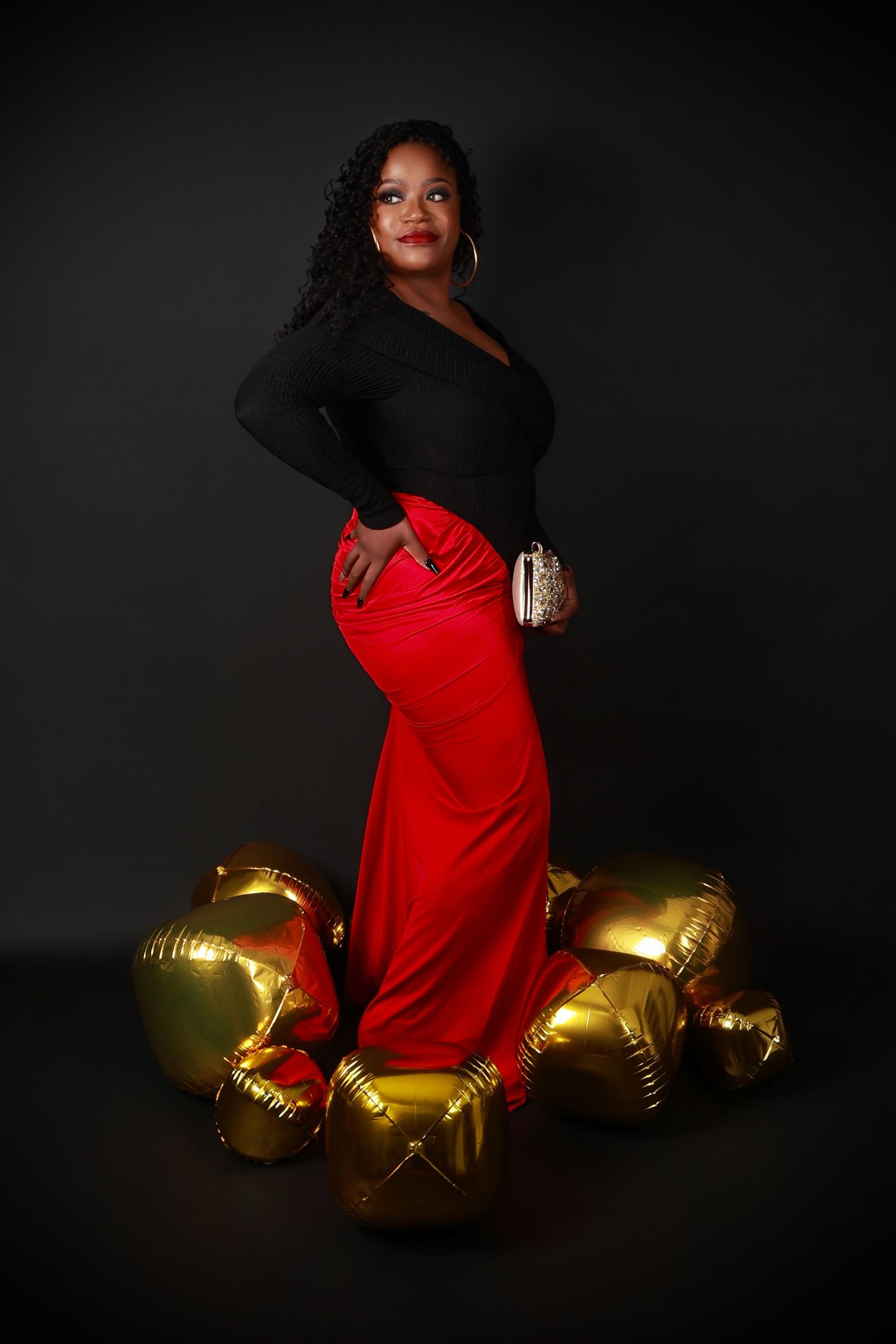 Woman in elegant black top and red maxi skirt posing with gold balloons on dark background.
