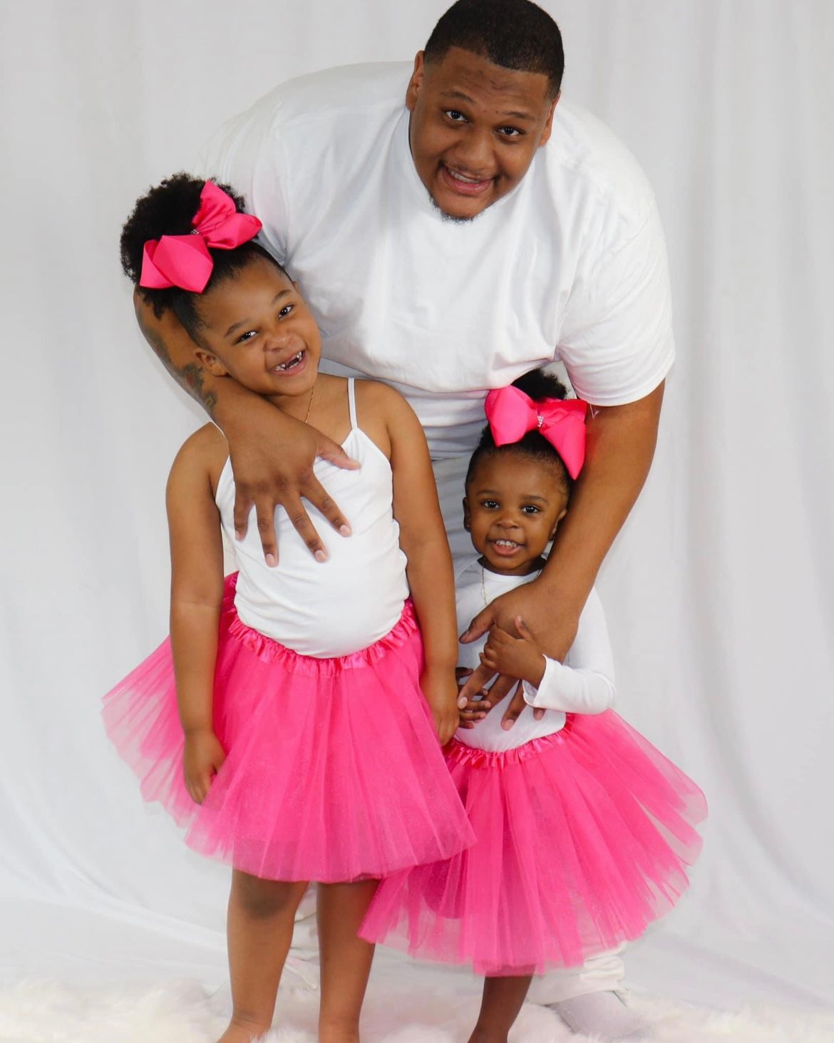 Father in white shirt poses with two young daughters in matching pink tutus and bows.