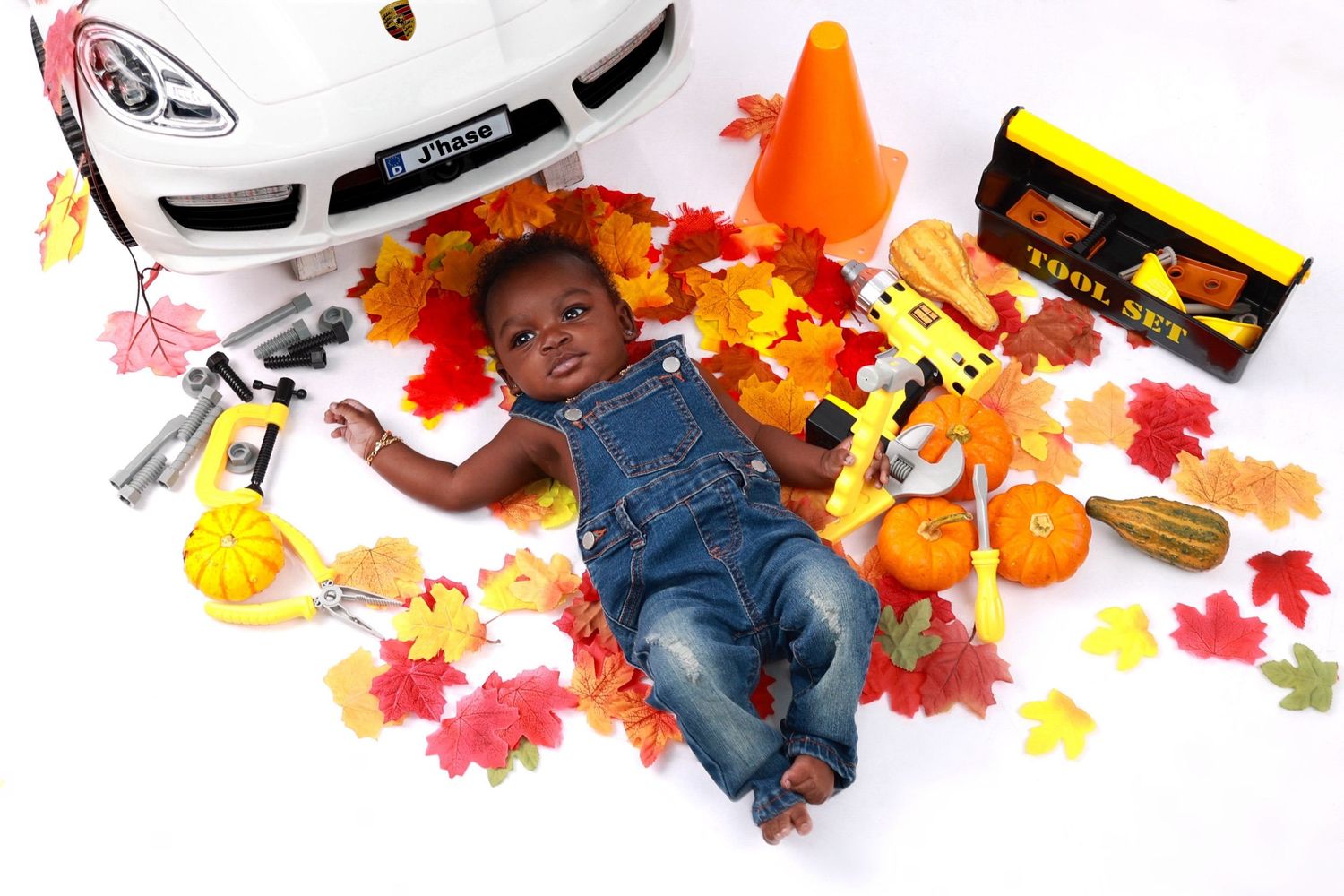 Baby boy in denim overalls lying among autumn leaves and toy tools beside a white car.