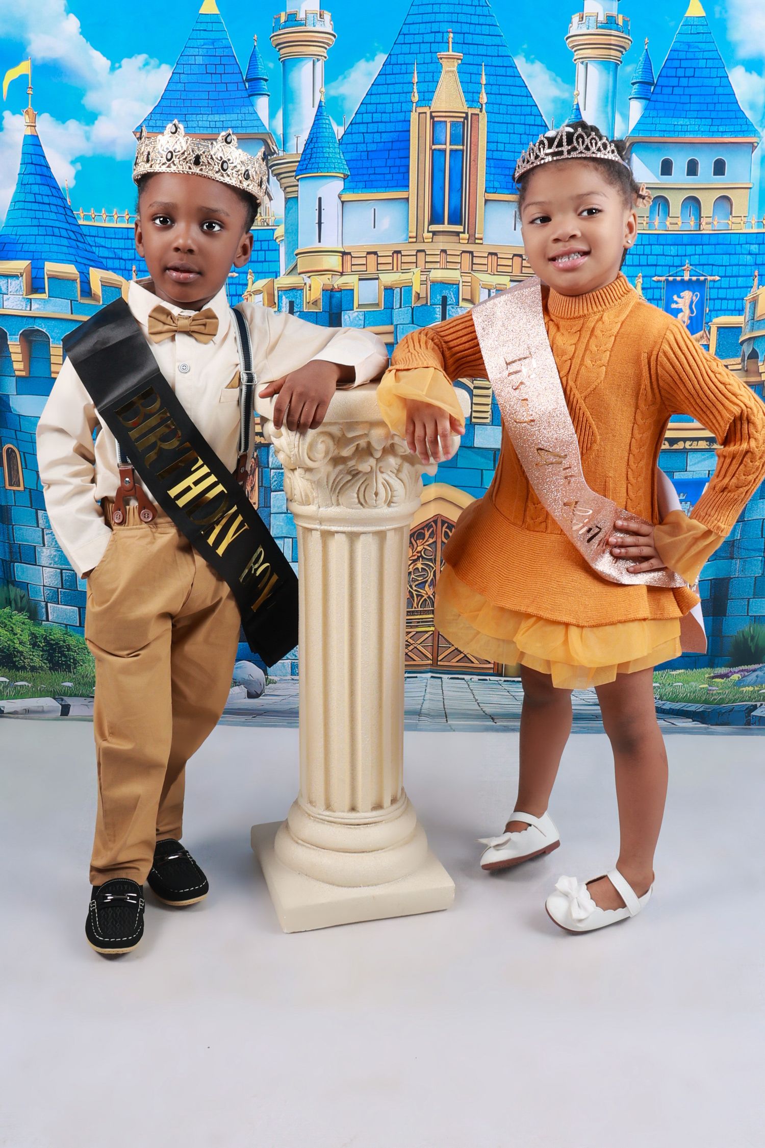 Two young children dressed as royalty pose in front of a castle backdrop wearing crowns and sashes.