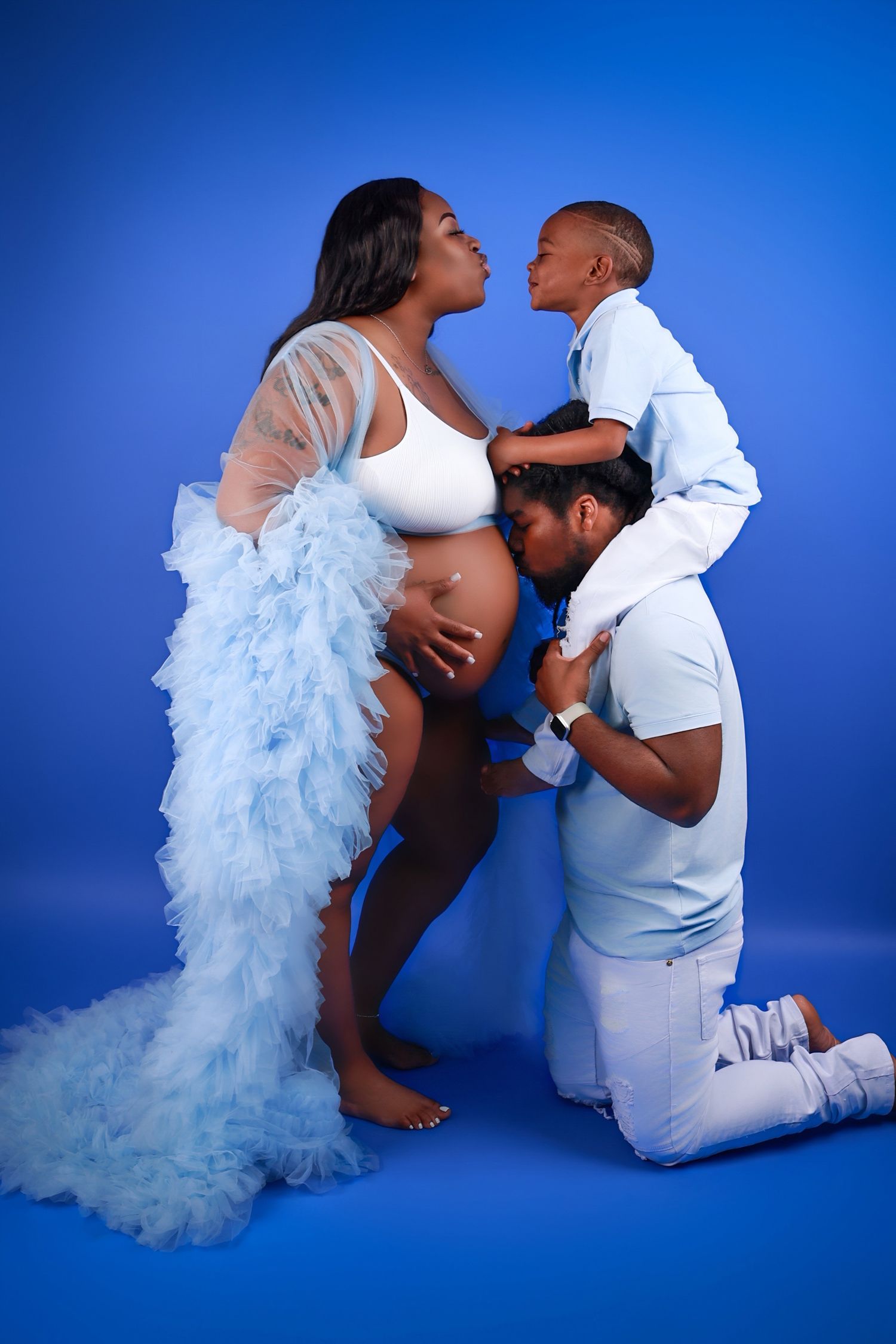 Pregnant woman in blue ruffled gown with partner kissing belly and child sharing a kiss above.