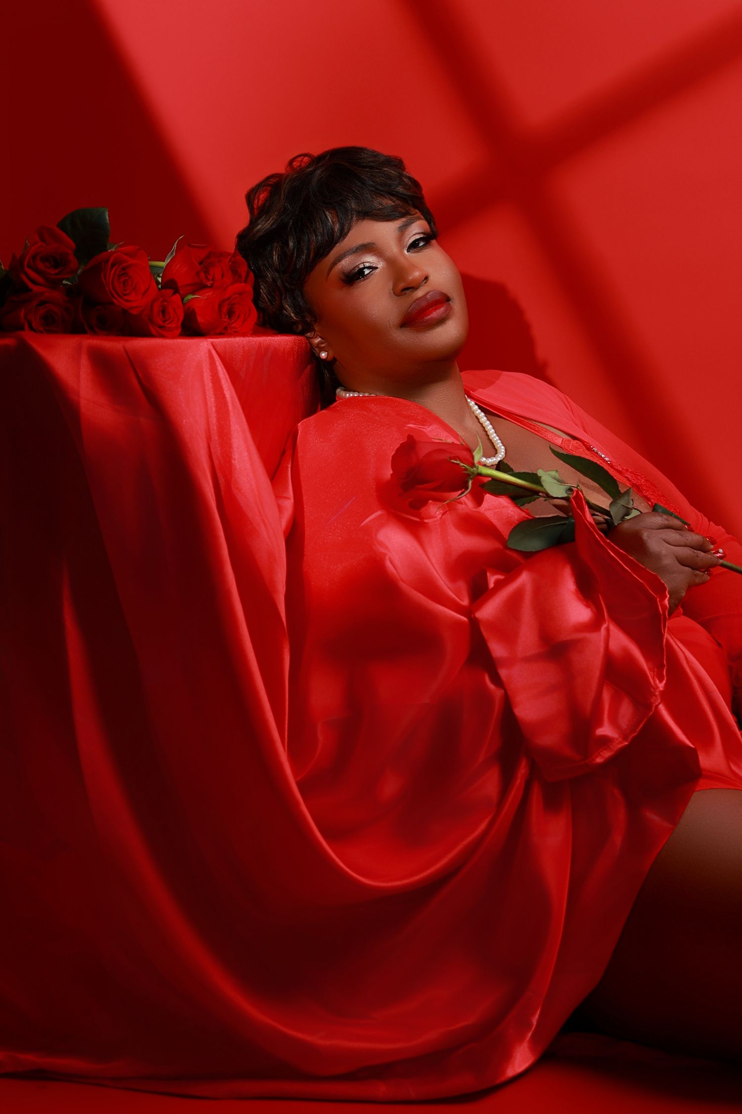 Woman in red satin robe holds a rose while posing against a bold red background.