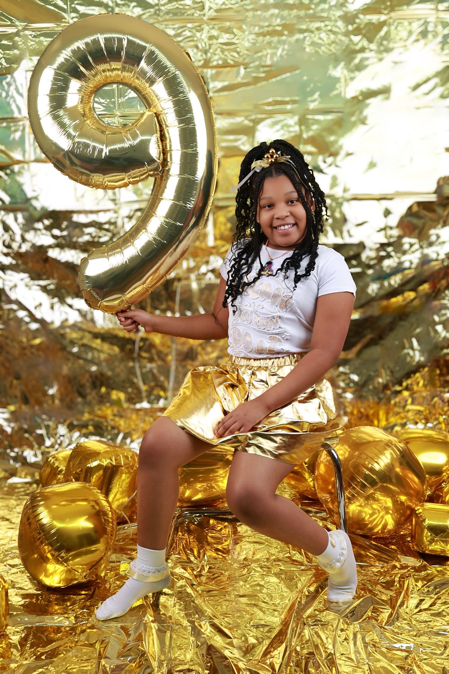 Girl celebrating 9th birthday in gold outfit holding number 9 balloon against gold backdrop.