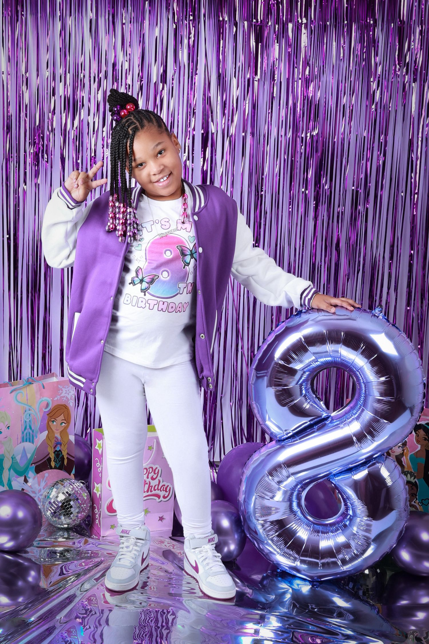 Girl celebrating 8th birthday in purple varsity jacket with silver balloon number 8 and purple decor.