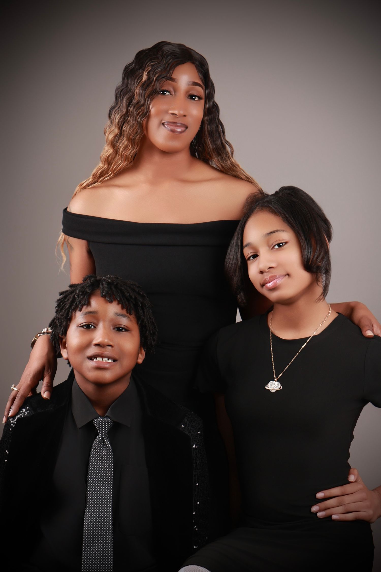 A woman in a black off-shoulder dress poses with two children in matching black outfits in a studio.