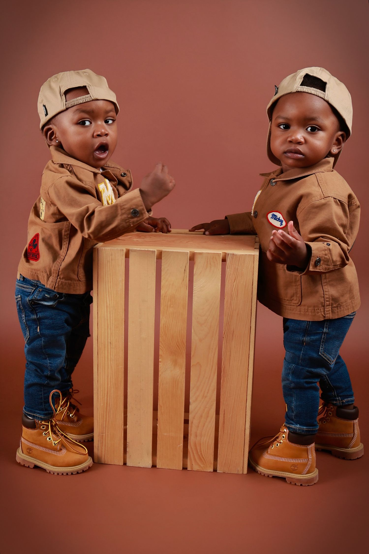 Two adorable Black toddler twins in matching tan jackets, jeans, and boots pose with a wooden crate.