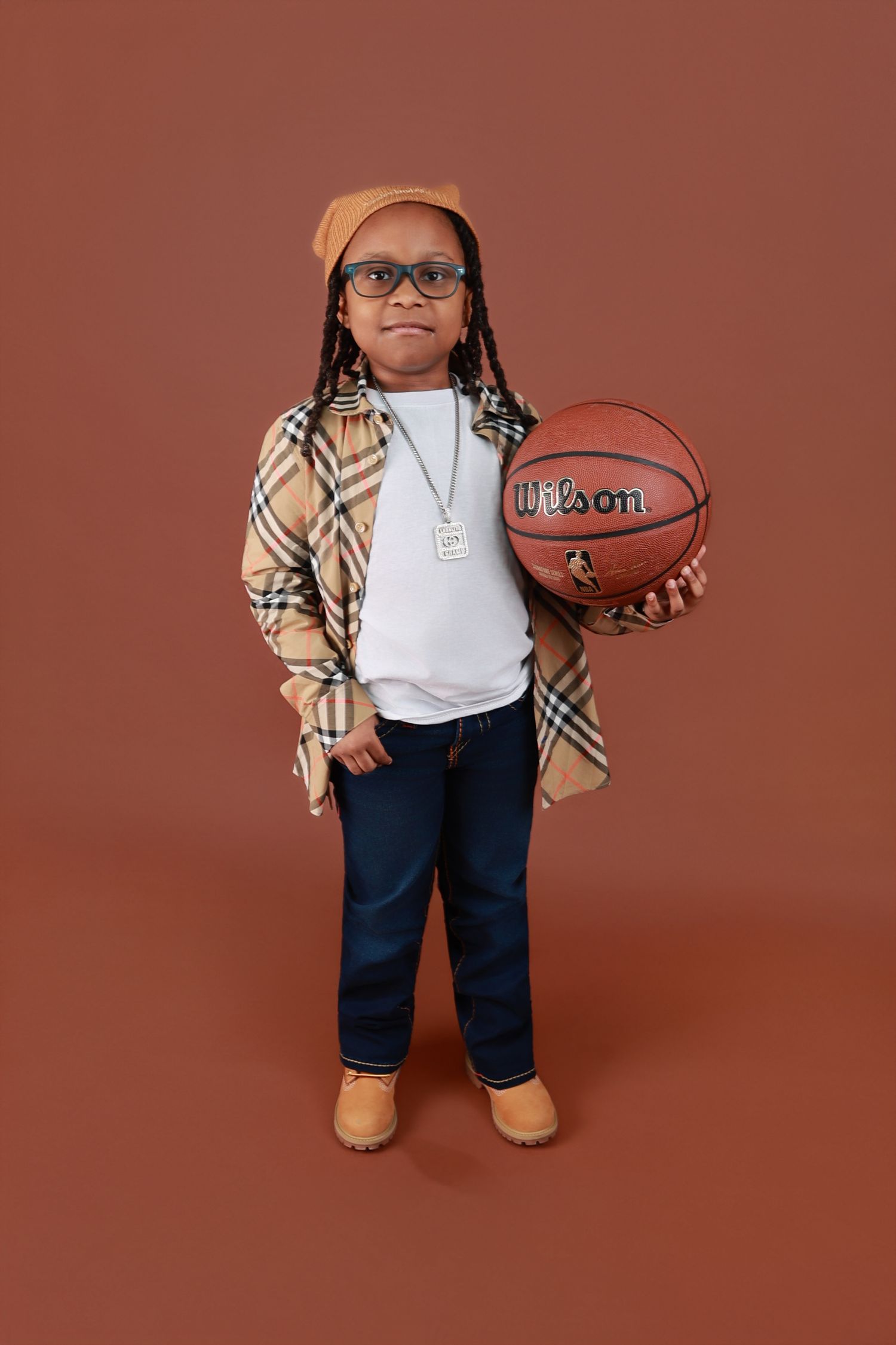Young boy in plaid jacket holding Wilson basketball on brown background.