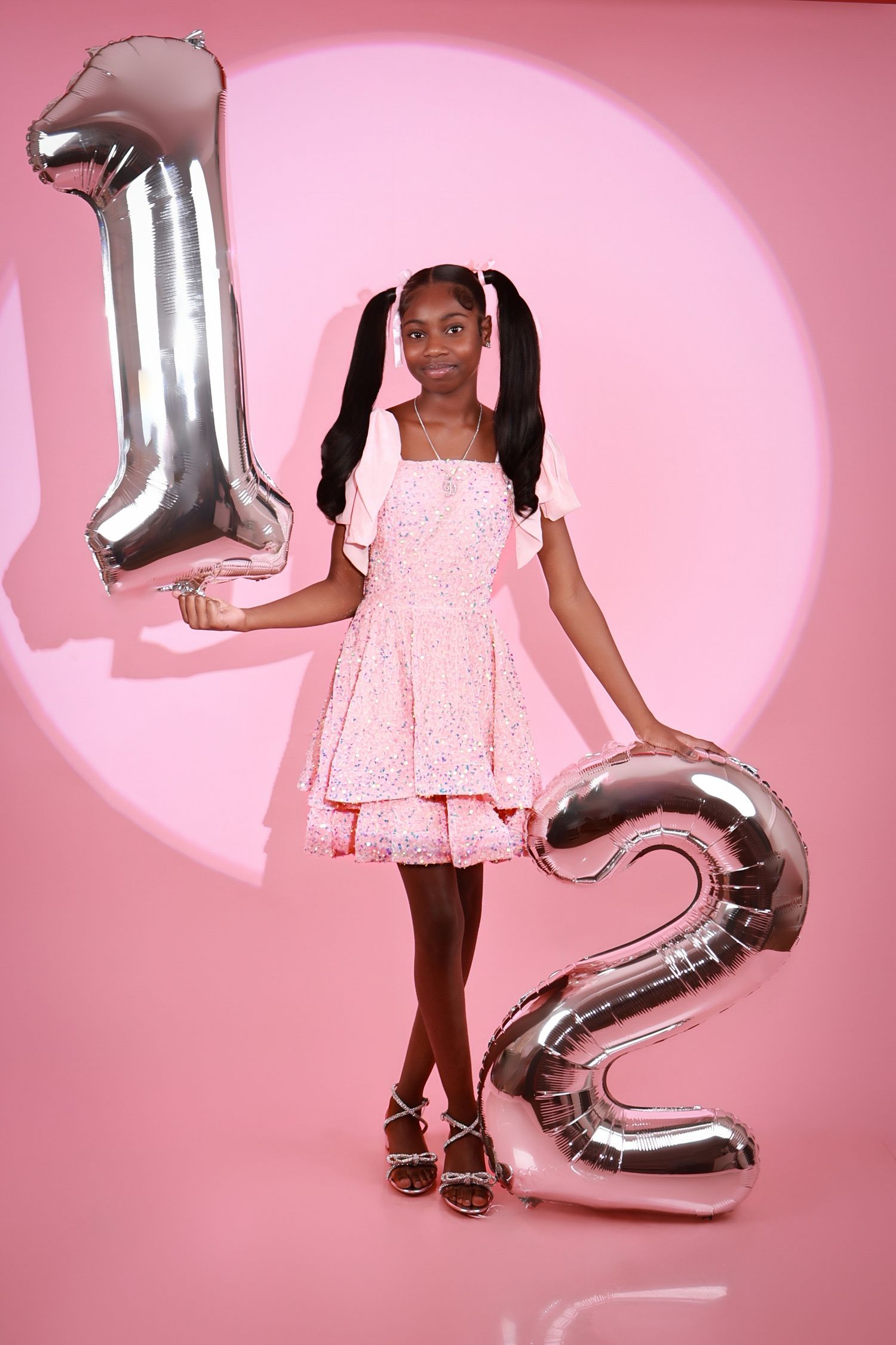 Young girl in pink dress holds large silver number 12 balloons celebrating her 12th birthday against a pink background.