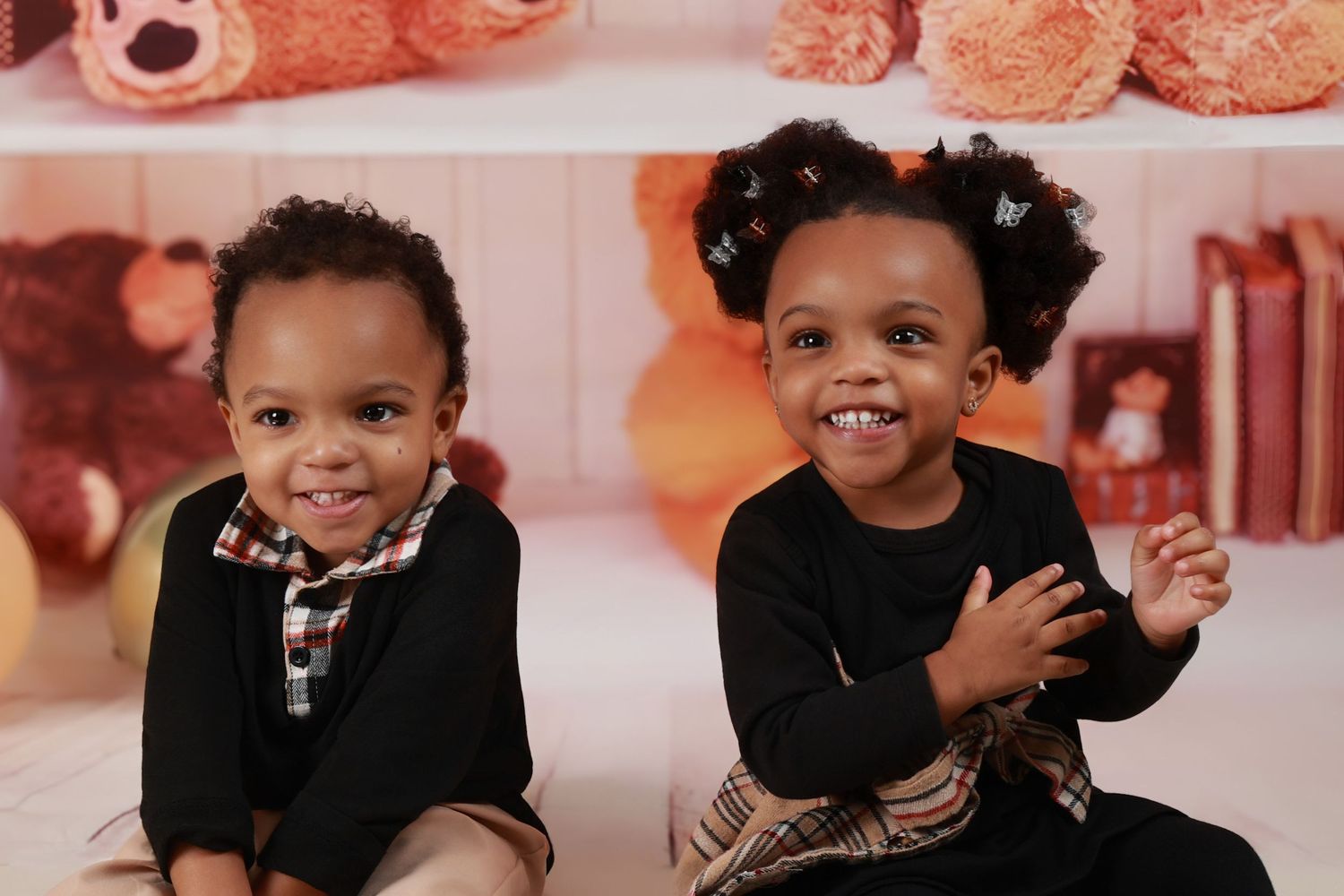 Two adorable young Black children smile together in front of a warm teddy bear backdrop.