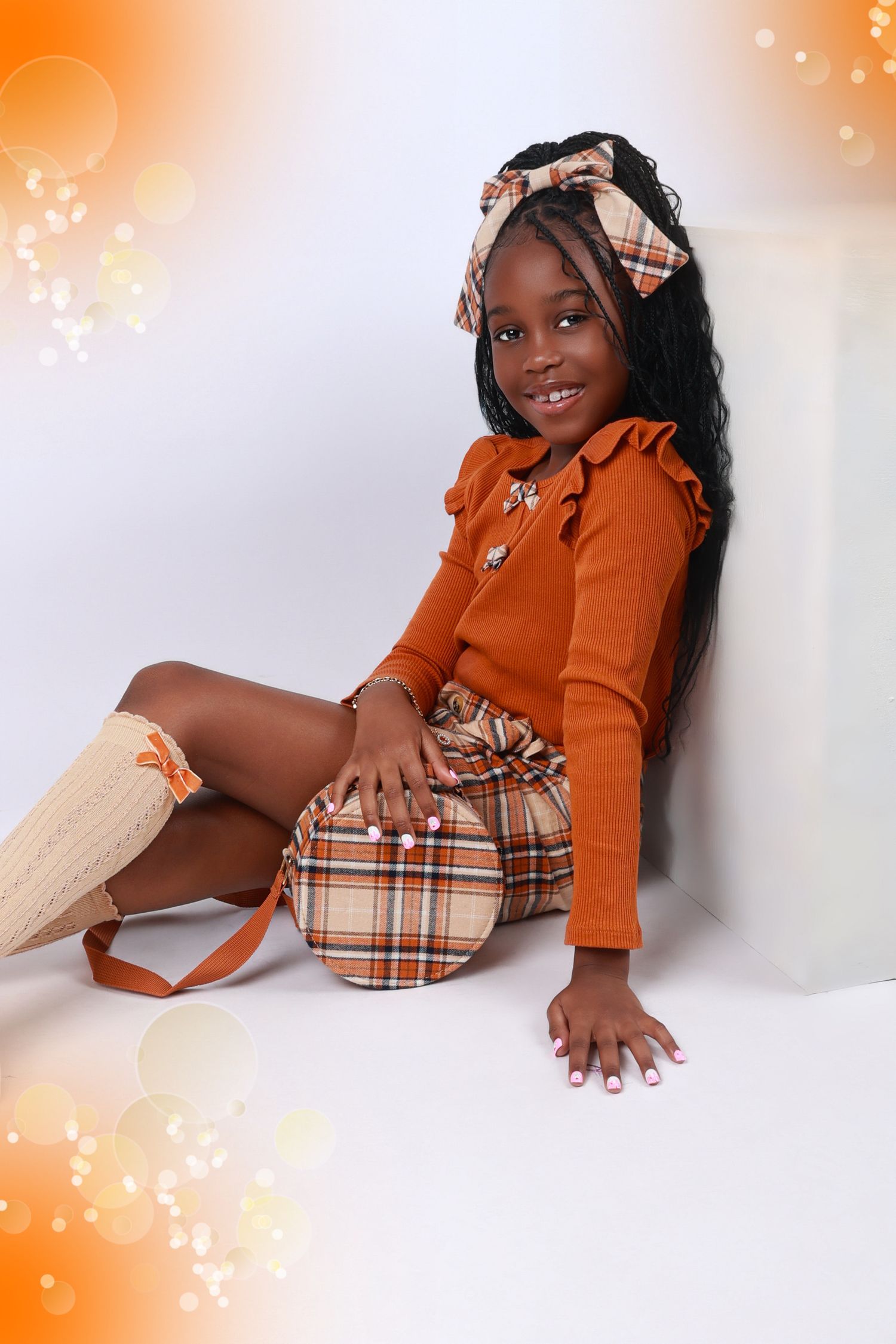 Young girl in orange top and plaid skirt with matching bow headband holding a purse in a studio.
