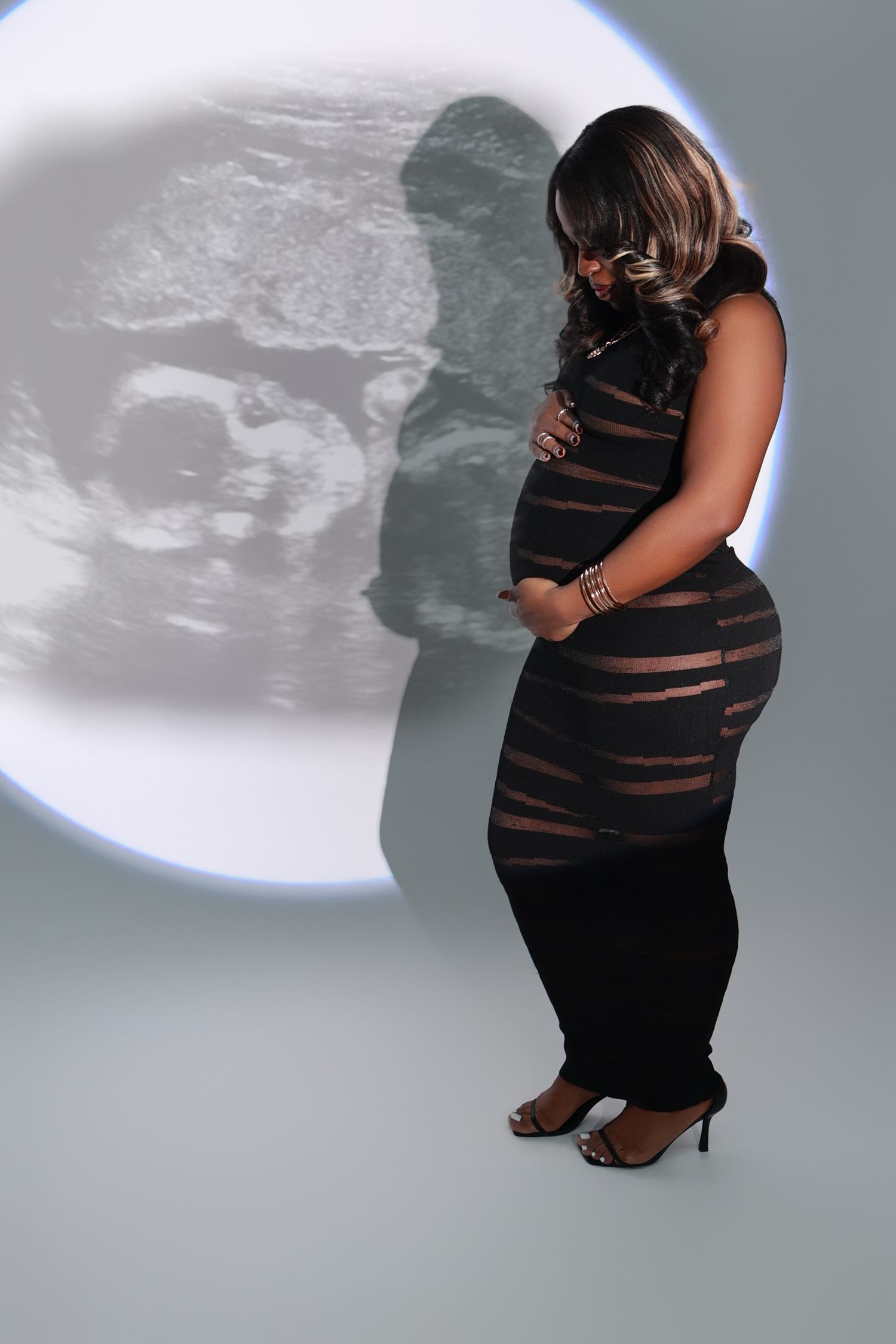 Pregnant woman in black dress cradling baby bump with ultrasound image projected behind her.