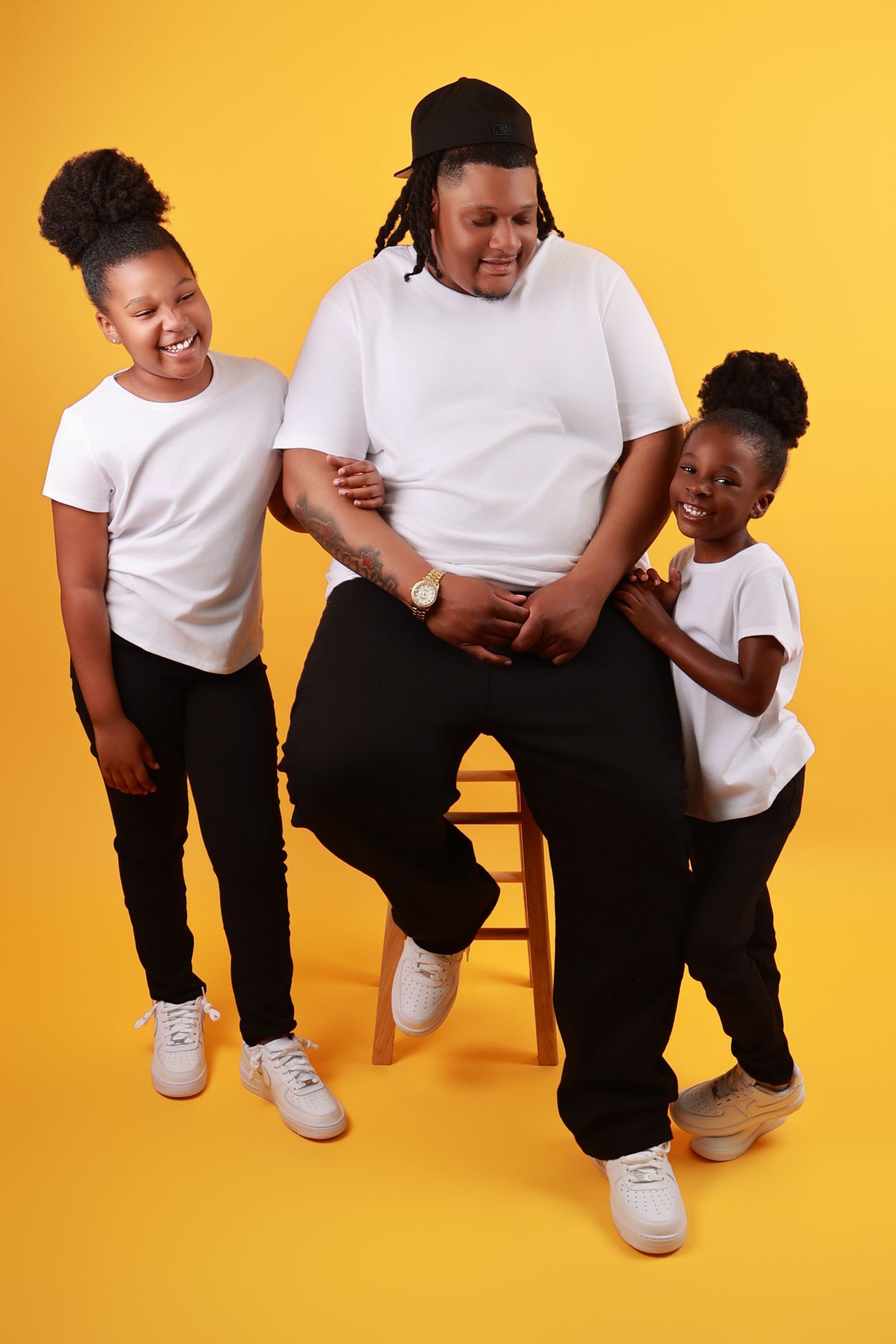 Father sits on stool with two daughters in matching white tees and black pants on yellow background.