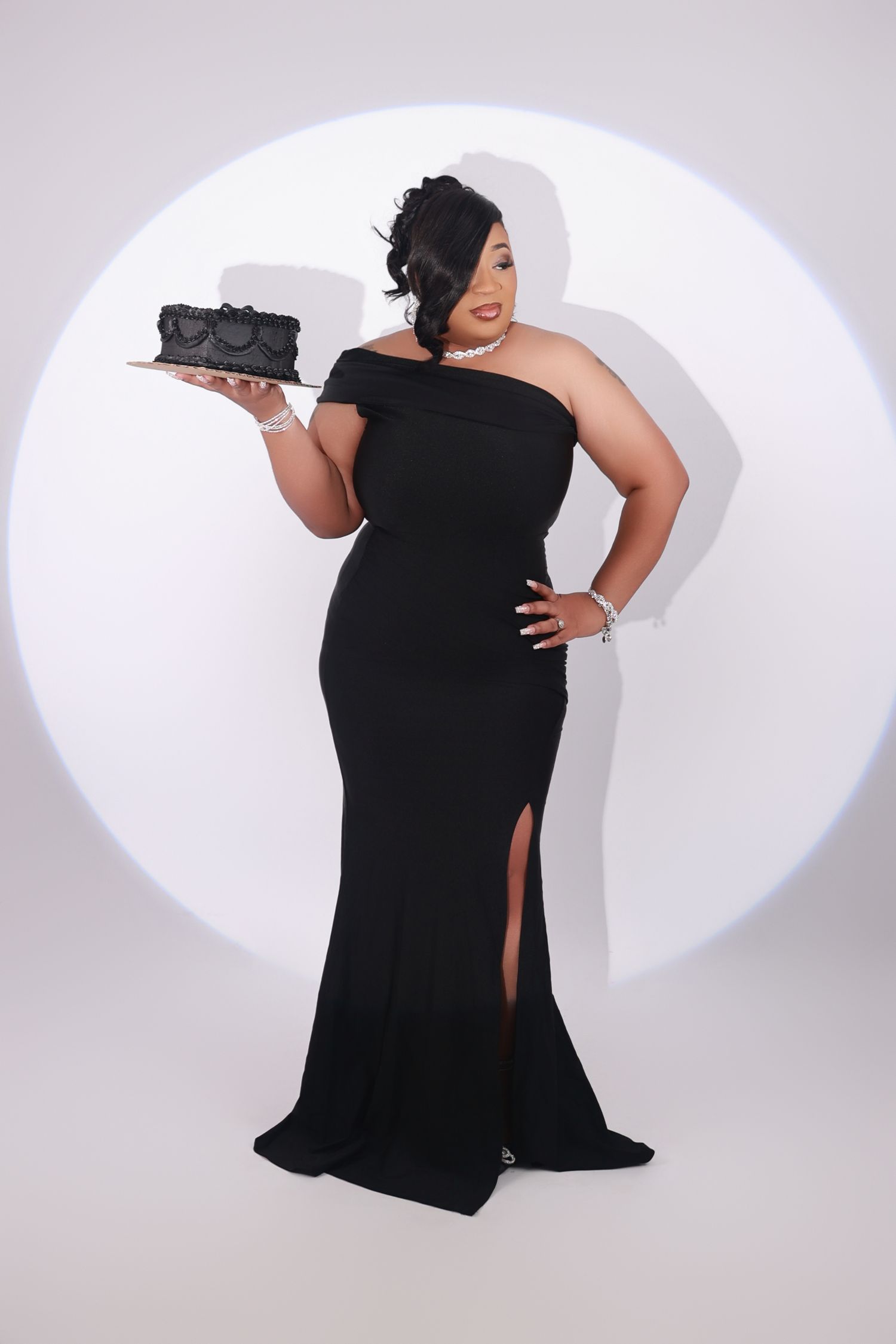 Woman in black off-shoulder gown holding decorative cake on serving tray in spotlight.