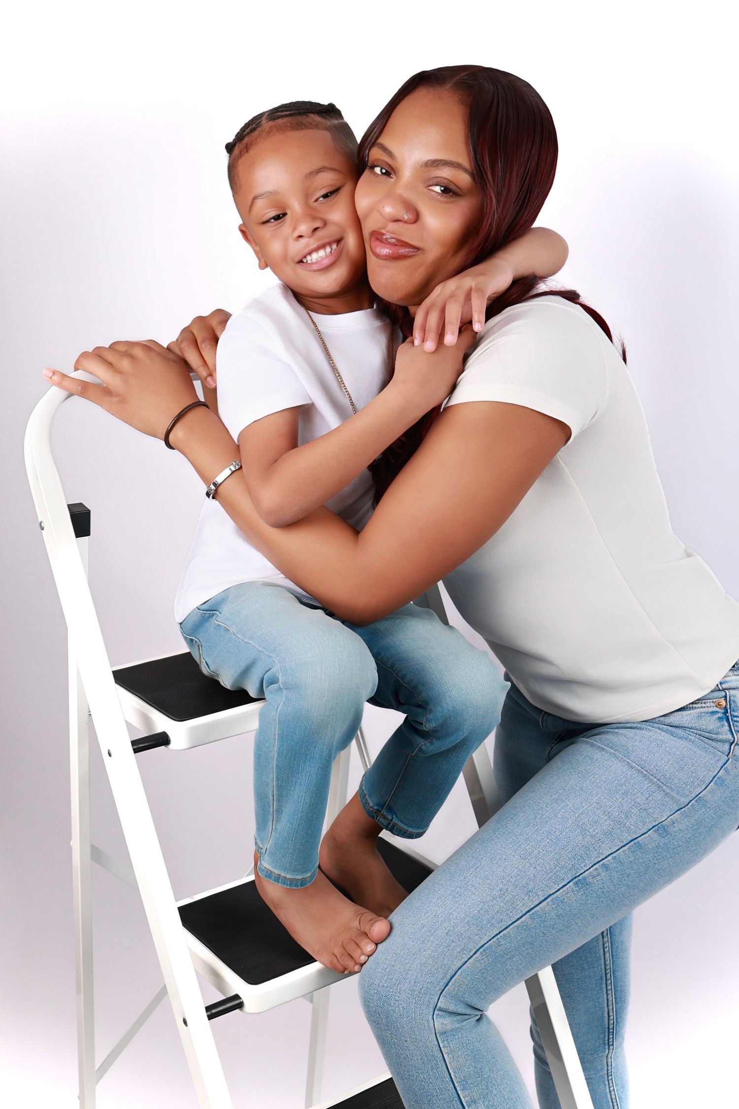 A young girl sits on a ladder hugging a woman in matching white tops and jeans in a studio portrait.