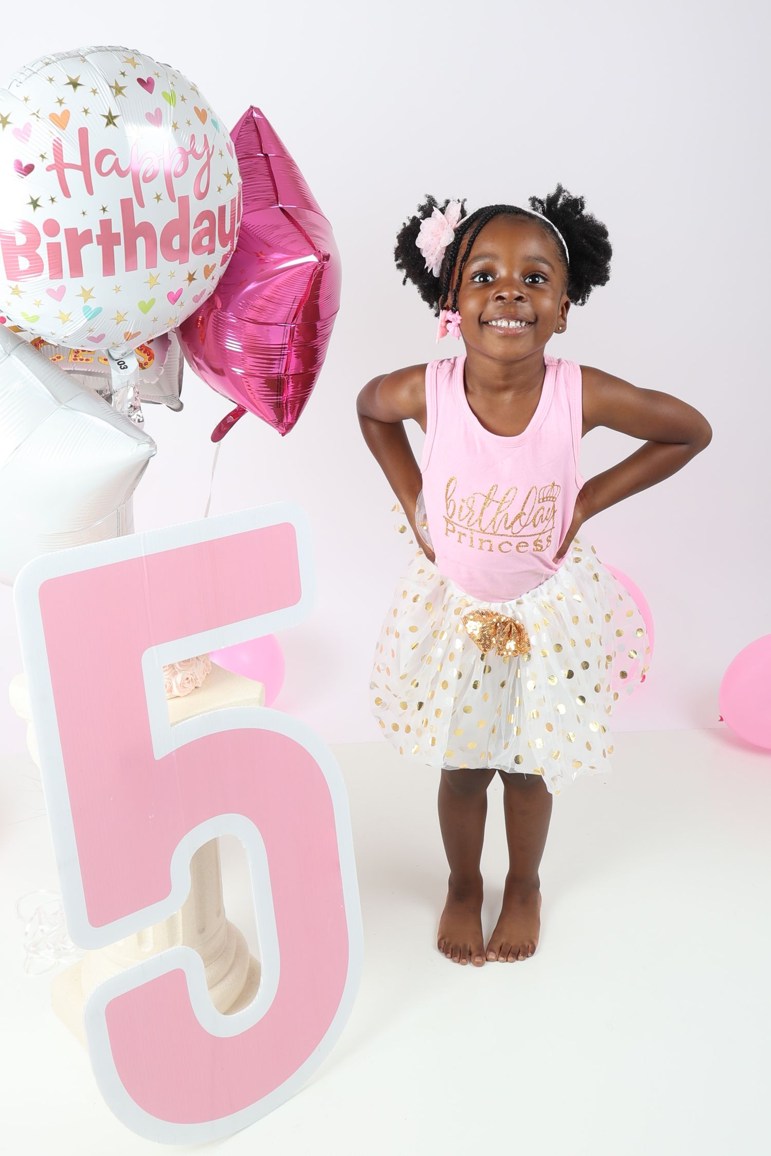 Happy girl in pink birthday princess outfit stands beside a large number 5 balloon display.