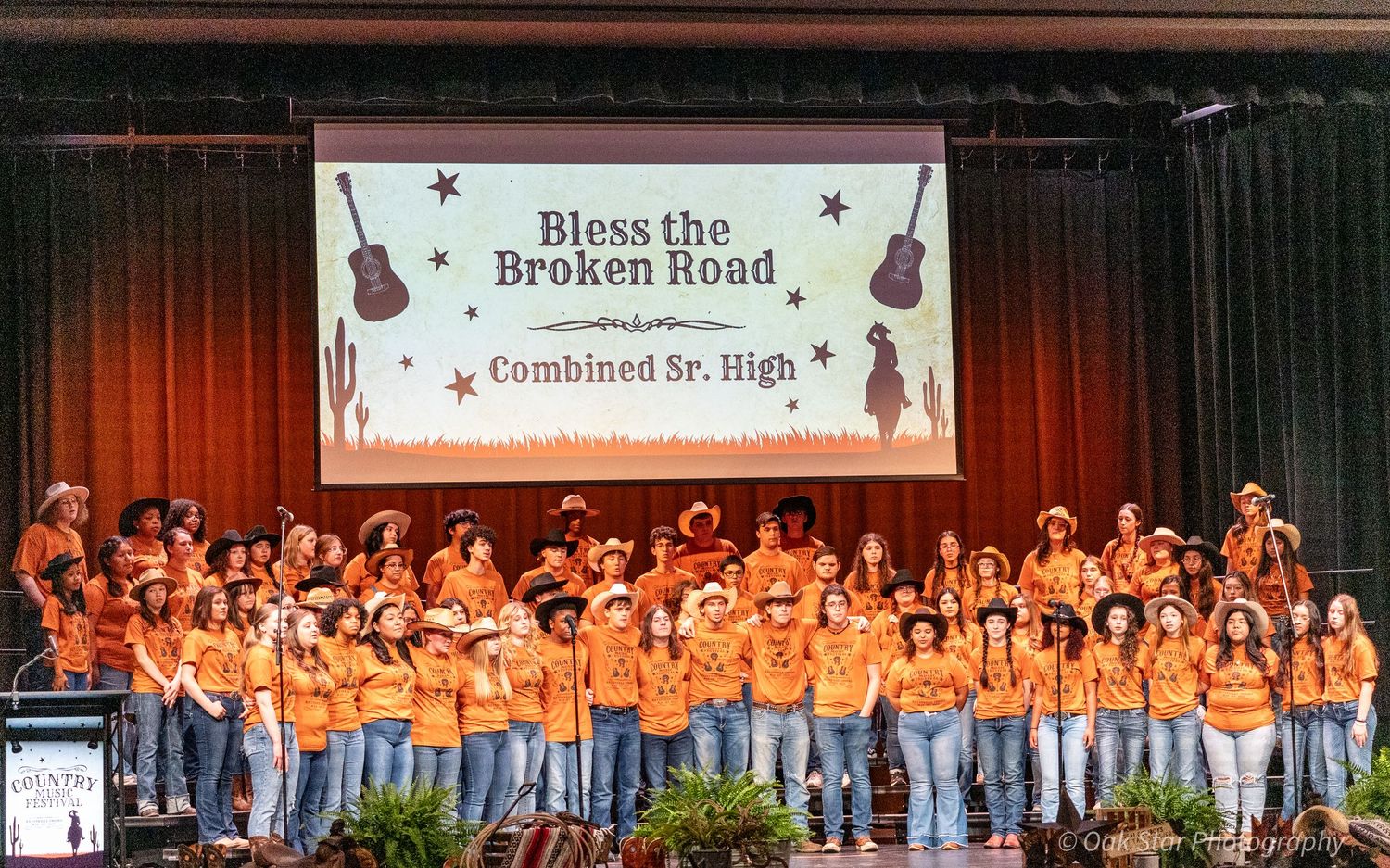 Choir group in matching orange shirts singing on stage with a large banner projected overhead.
