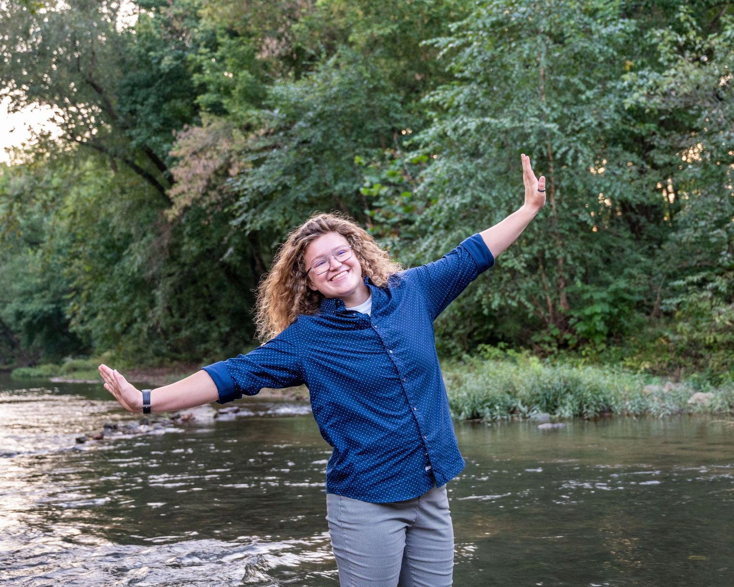 Outdoor senior portrait of a young lady standing at a river with trees in the background.