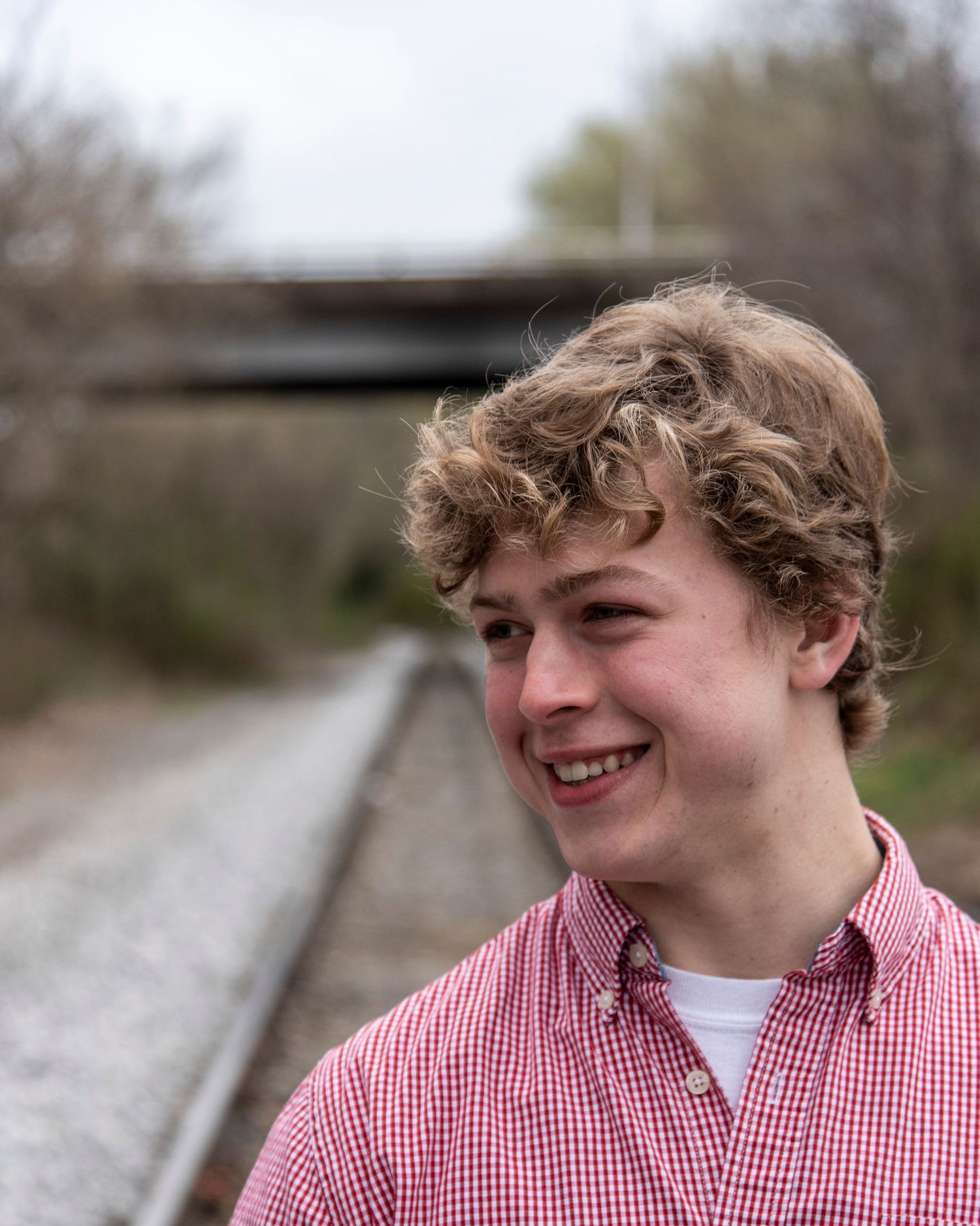 Outdoor senior portrait of a smiling young man standing by a railroad track with a bridge in the background.
