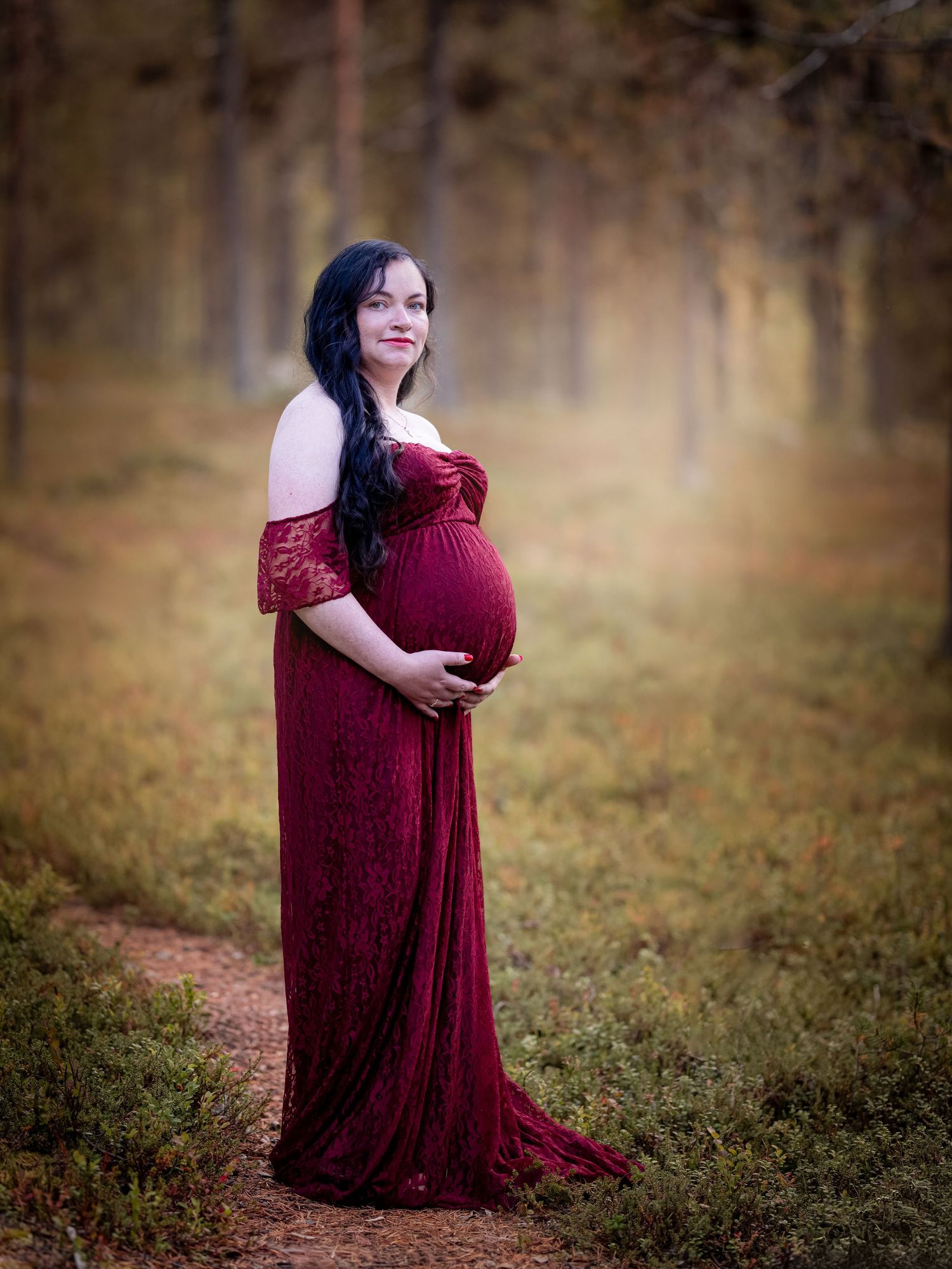 Person in a flowing burgundy dress poses in a wooded area with soft natural lighting and autumn tones.
