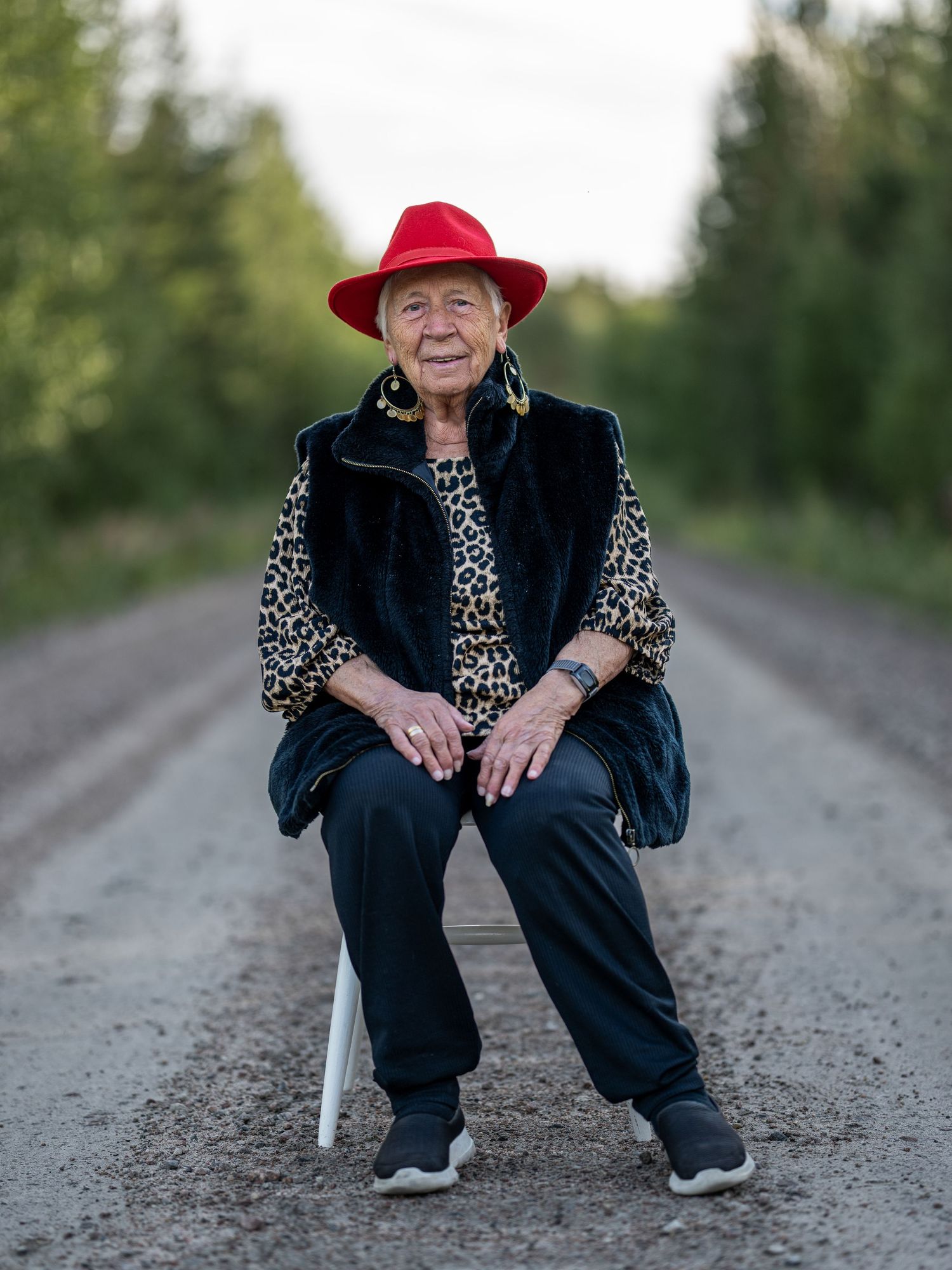 A person wearing black clothing and a red hat sits casually on a gravel road in a rural setting.