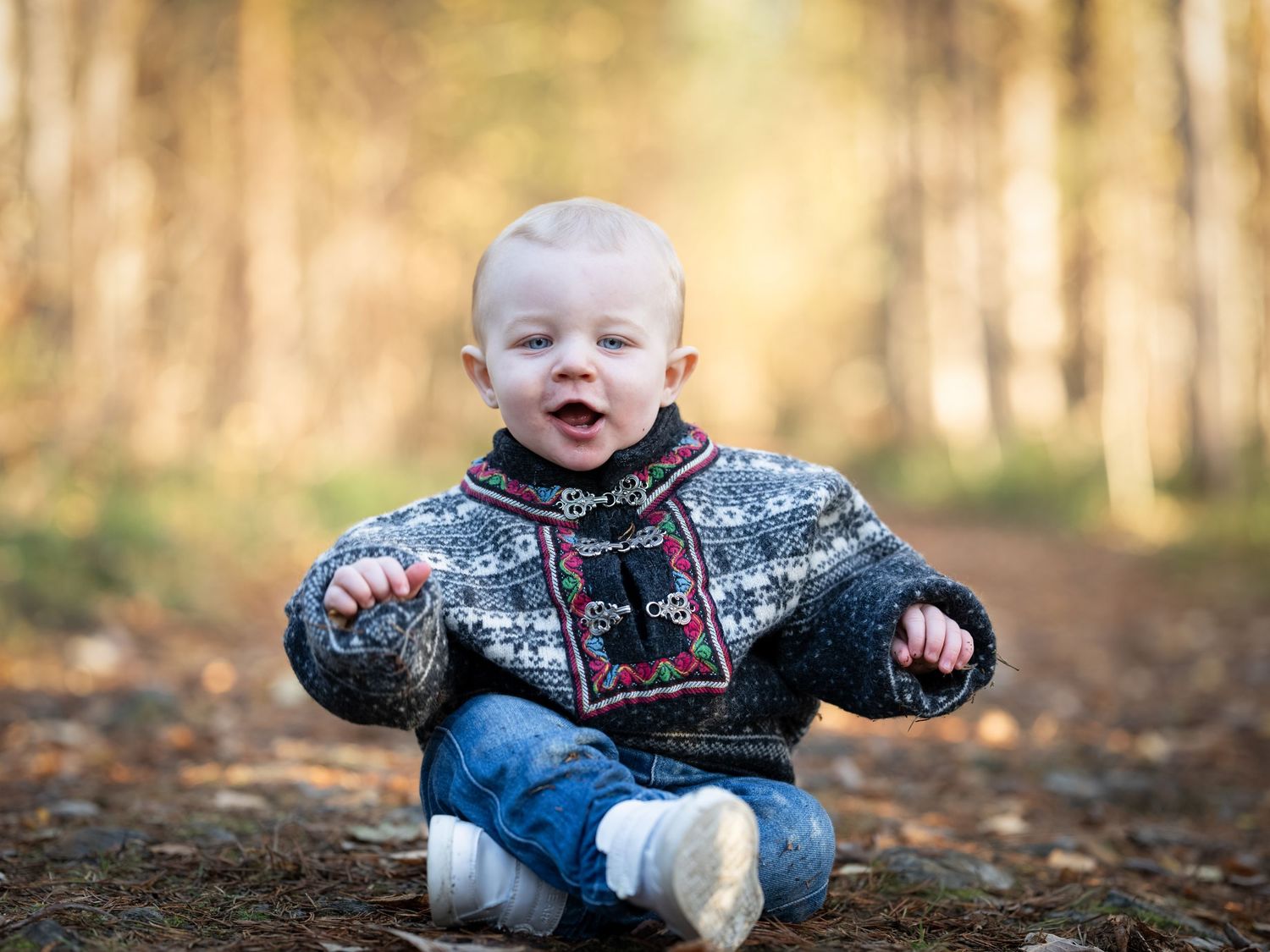 A small child sits on fallen autumn leaves wearing a dark patterned sweater and jeans in an outdoor setting.