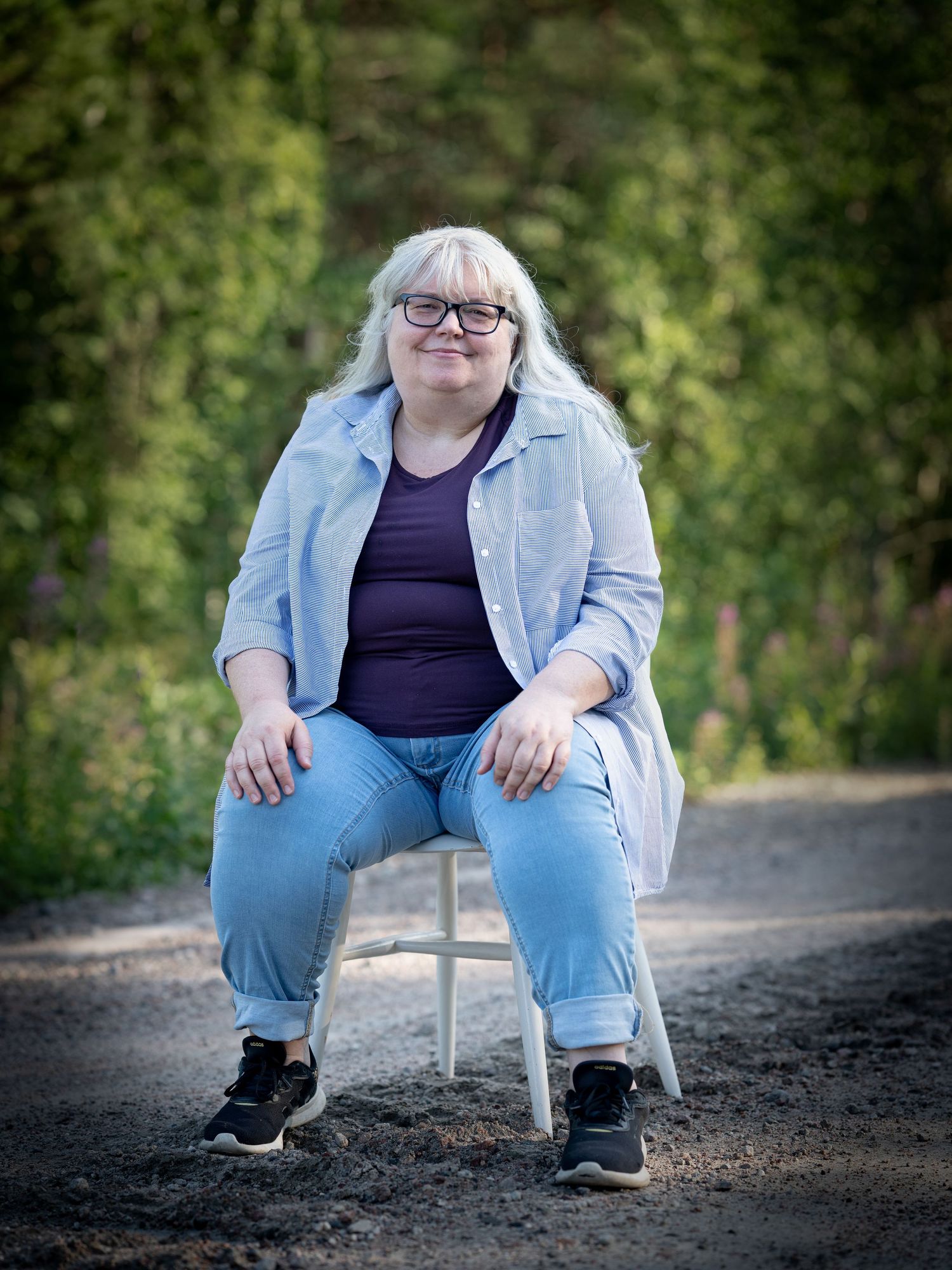 A person wearing jeans and a light denim jacket sits on a white folding chair outdoors against a blurred green background.