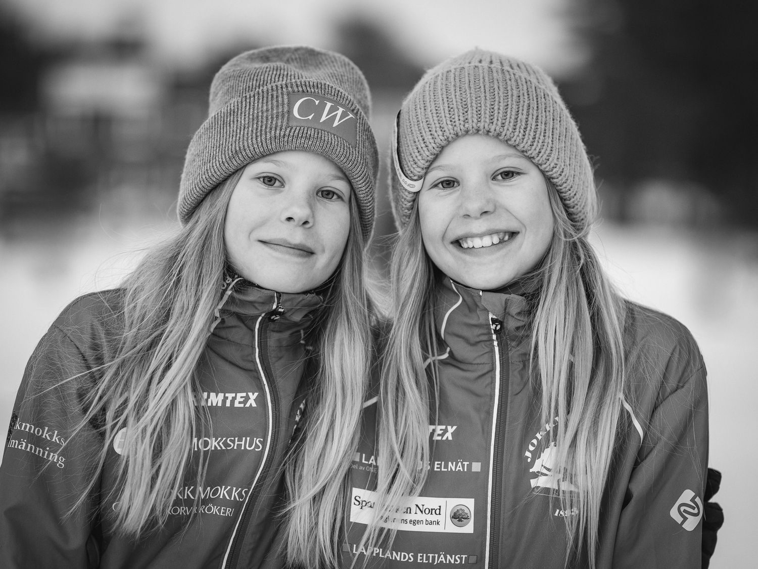 Two winter athletes wearing matching gray knit hats and team jackets pose together.