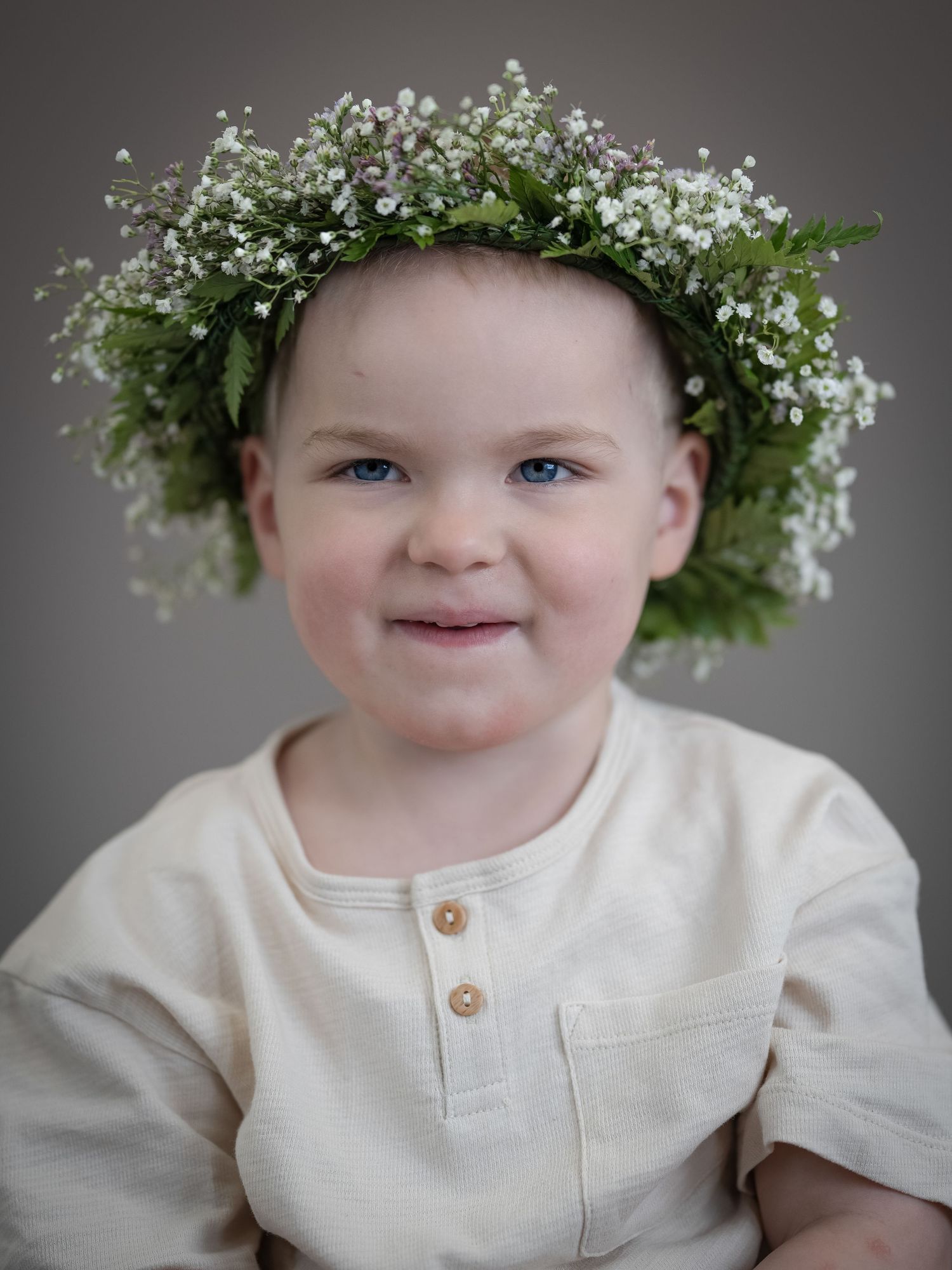 Ett porträtt med en delikat babyandning och en grönskande blomsterkrona mot en grå bakgrund.