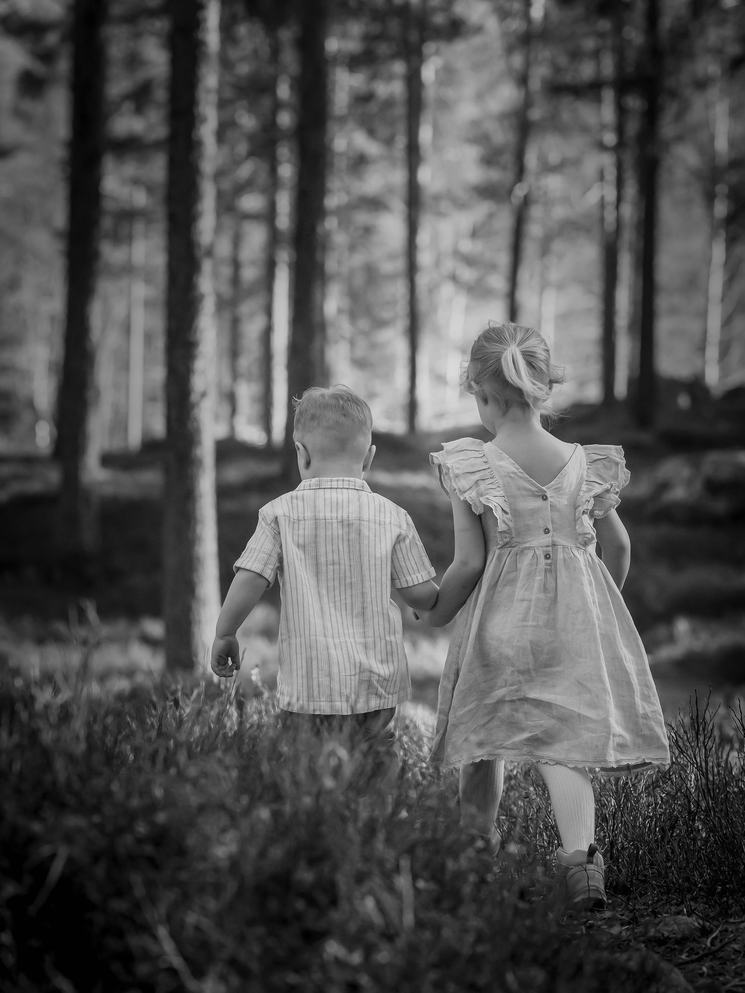 Black and white image of two small children walking hand in hand through a misty forest.