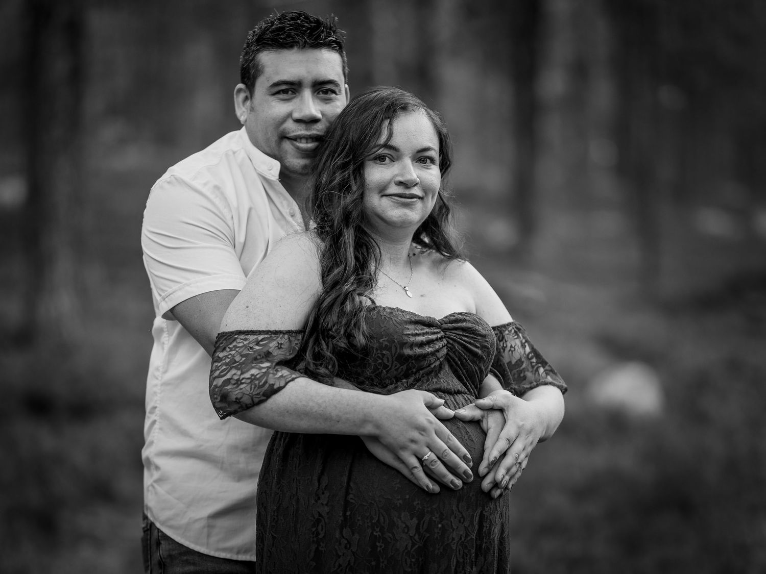 Black and white outdoor portrait of a couple embracing during a romantic maternity photoshoot in a forest setting.