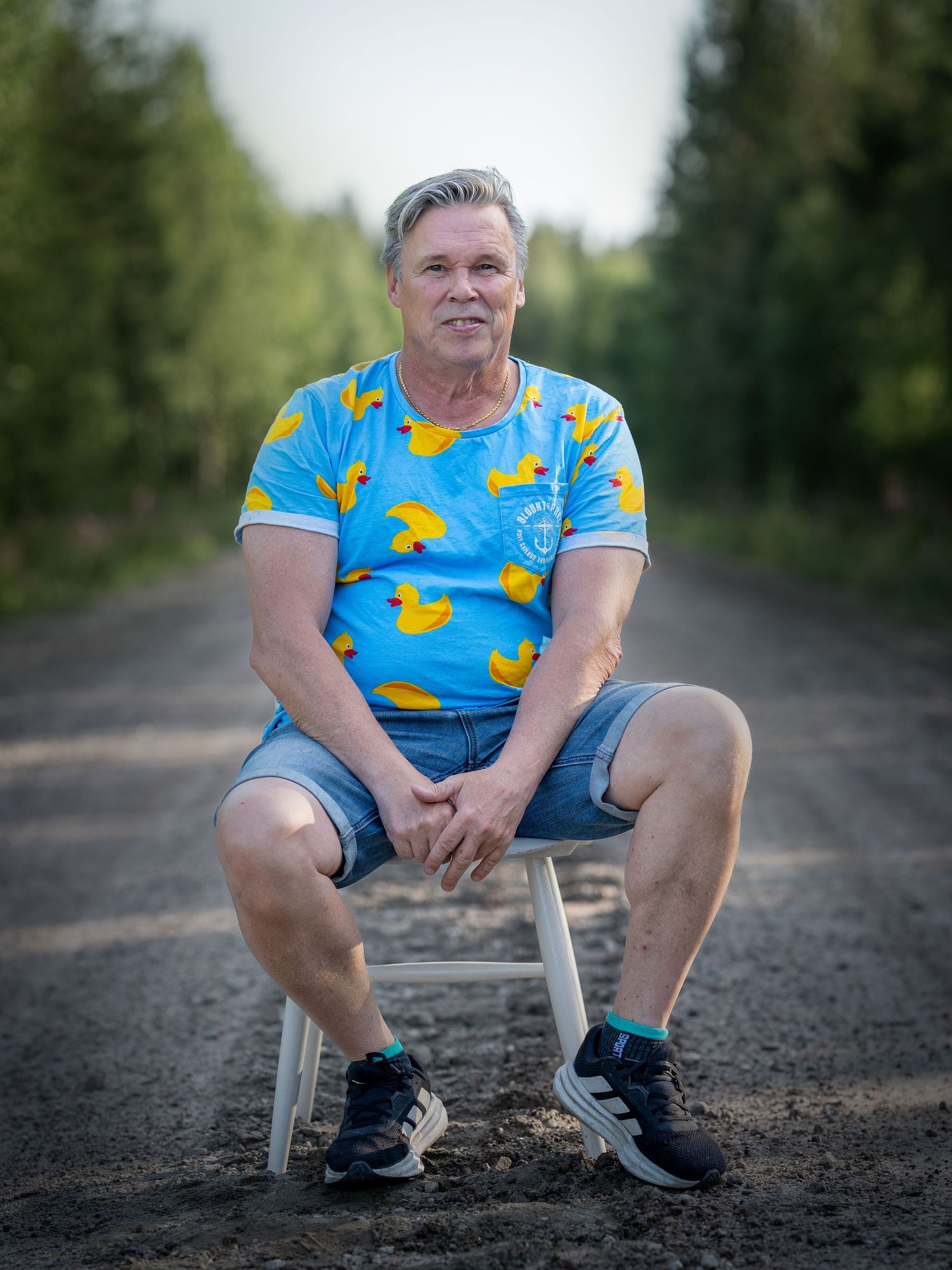 Person wearing a blue rubber duck print t-shirt sits on a small stool on a dirt path surrounded by greenery.