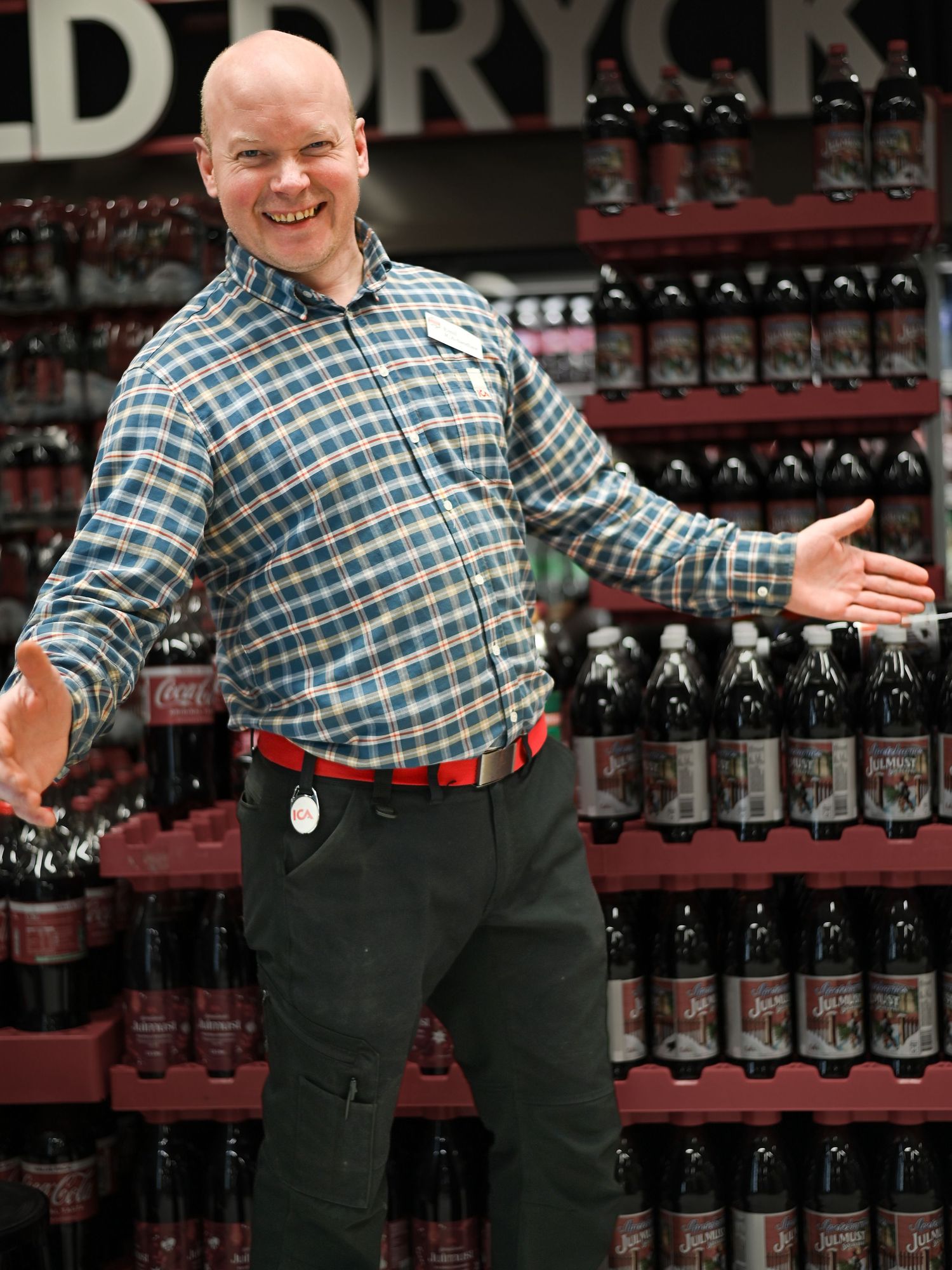 In a retail setting, a person in checkered shirt and dark pants gestures near shelves stocked with wine bottles.