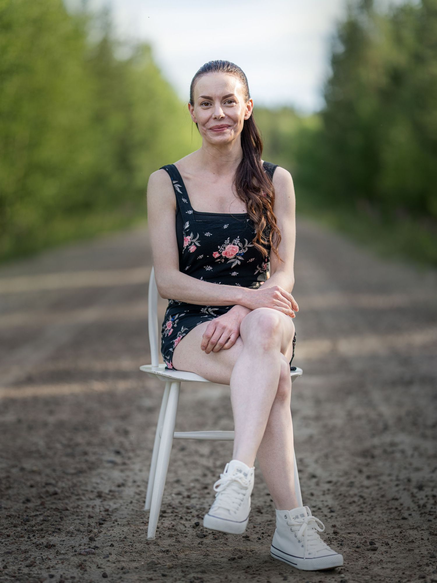 A person in a floral dress and sneakers sits on a white chair on a dirt path surrounded by greenery.