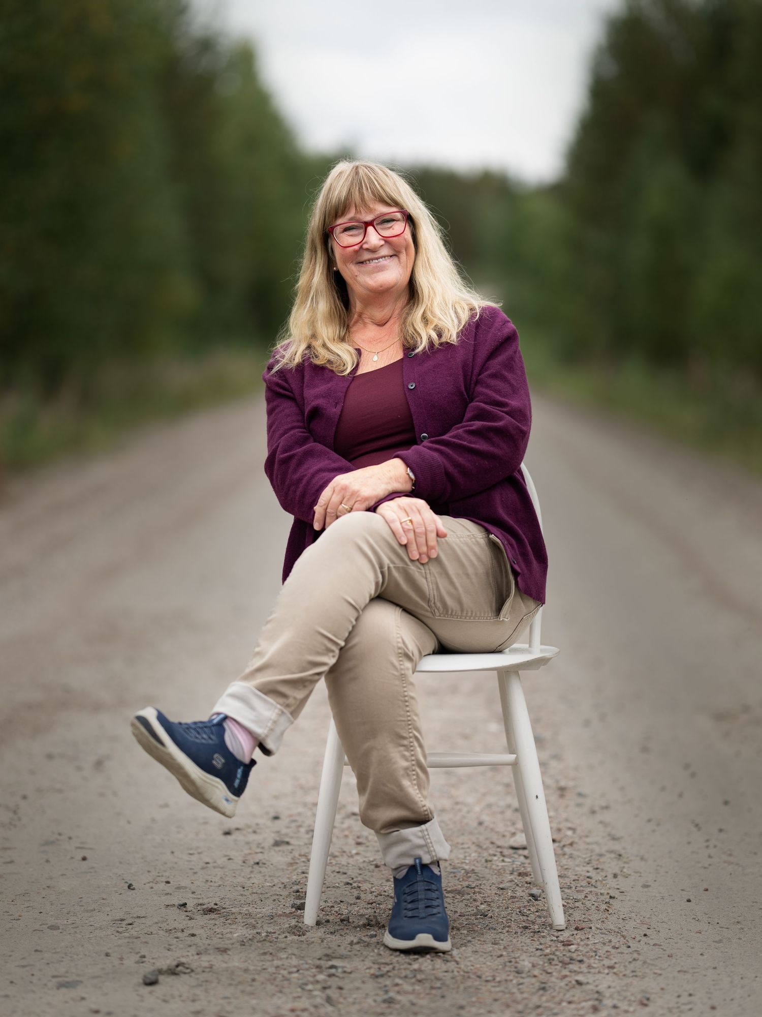 A casual portrait of someone sitting on a white stool along a tree-lined dirt road wearing a purple sweater.