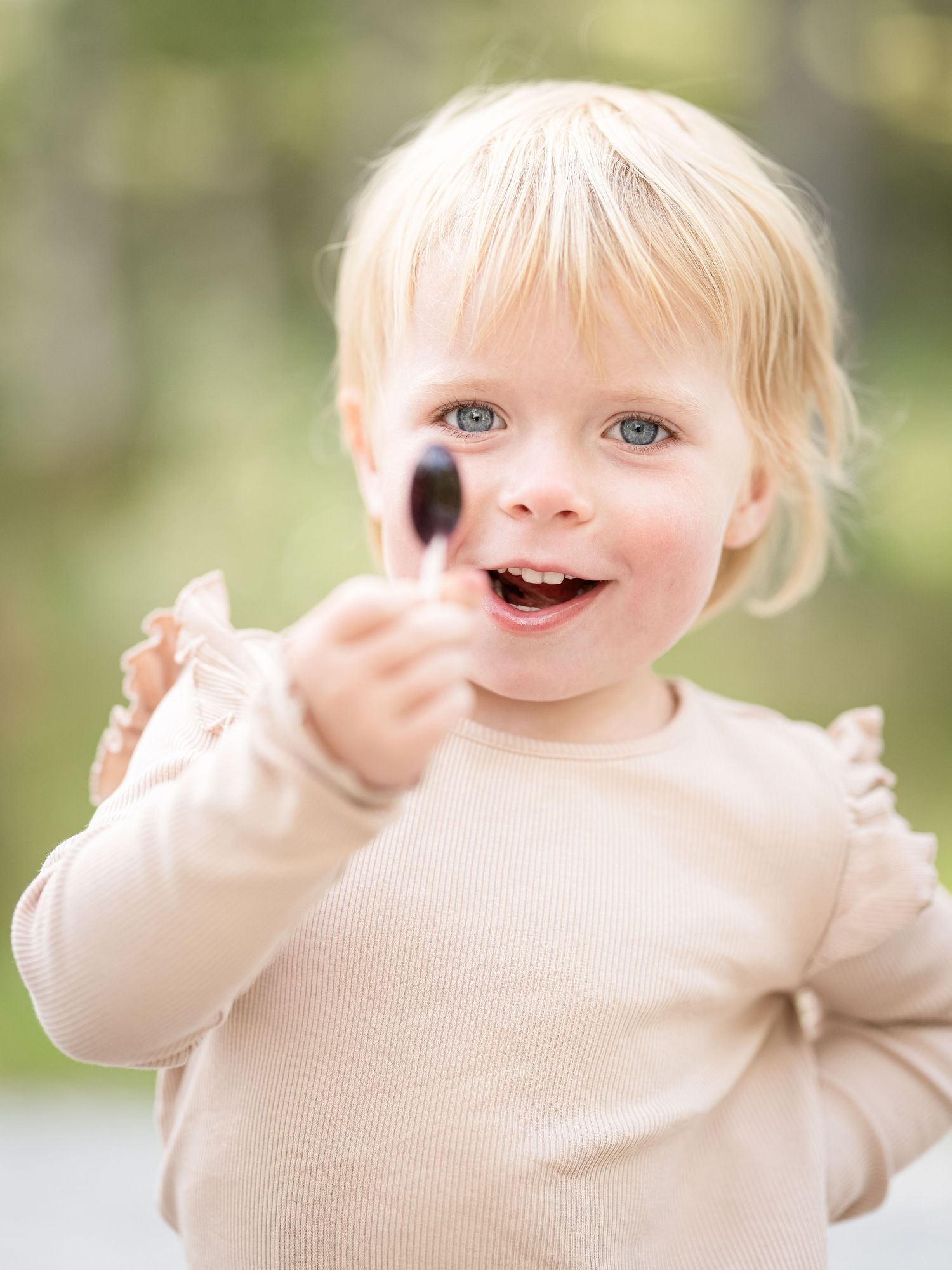 A toddler in a cream sweater makes silly faces and gestures while playing outdoors on a sunny day.