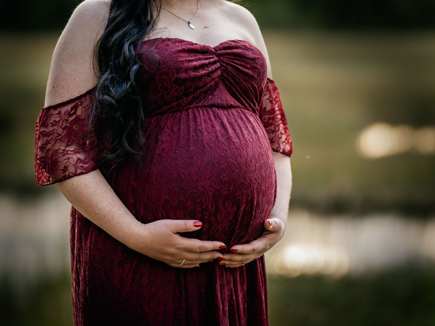 Maternity photo showing pregnant belly in burgundy lace off-shoulder dress against blurred outdoor background.