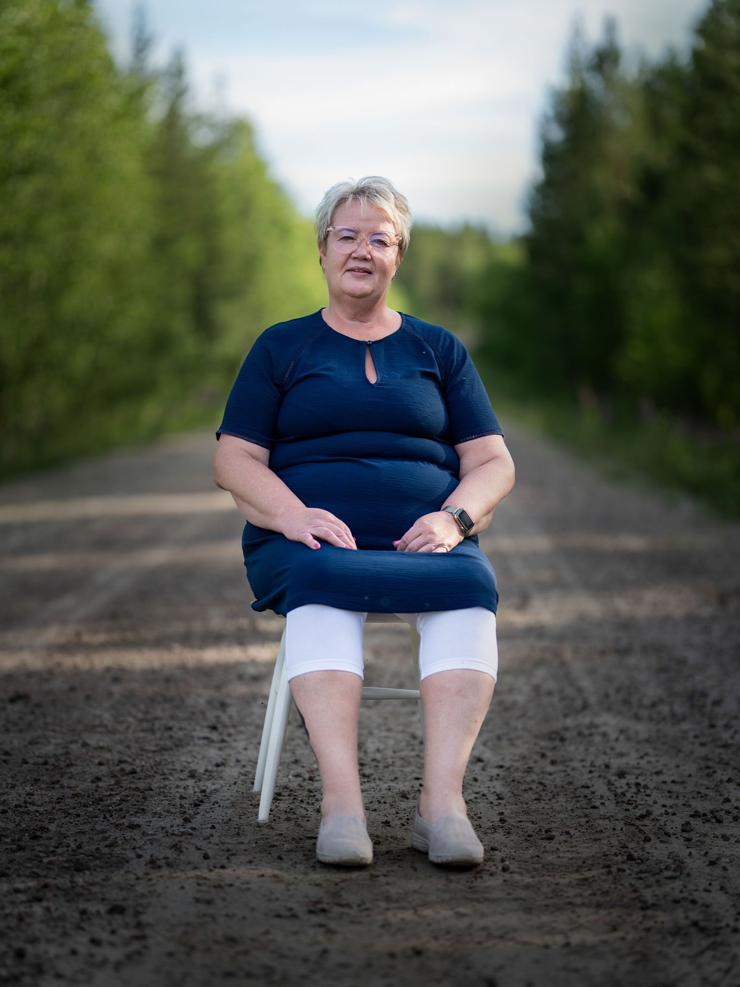 Person in a navy blue top and white pants sits on a white chair on a dirt path surrounded by green trees.