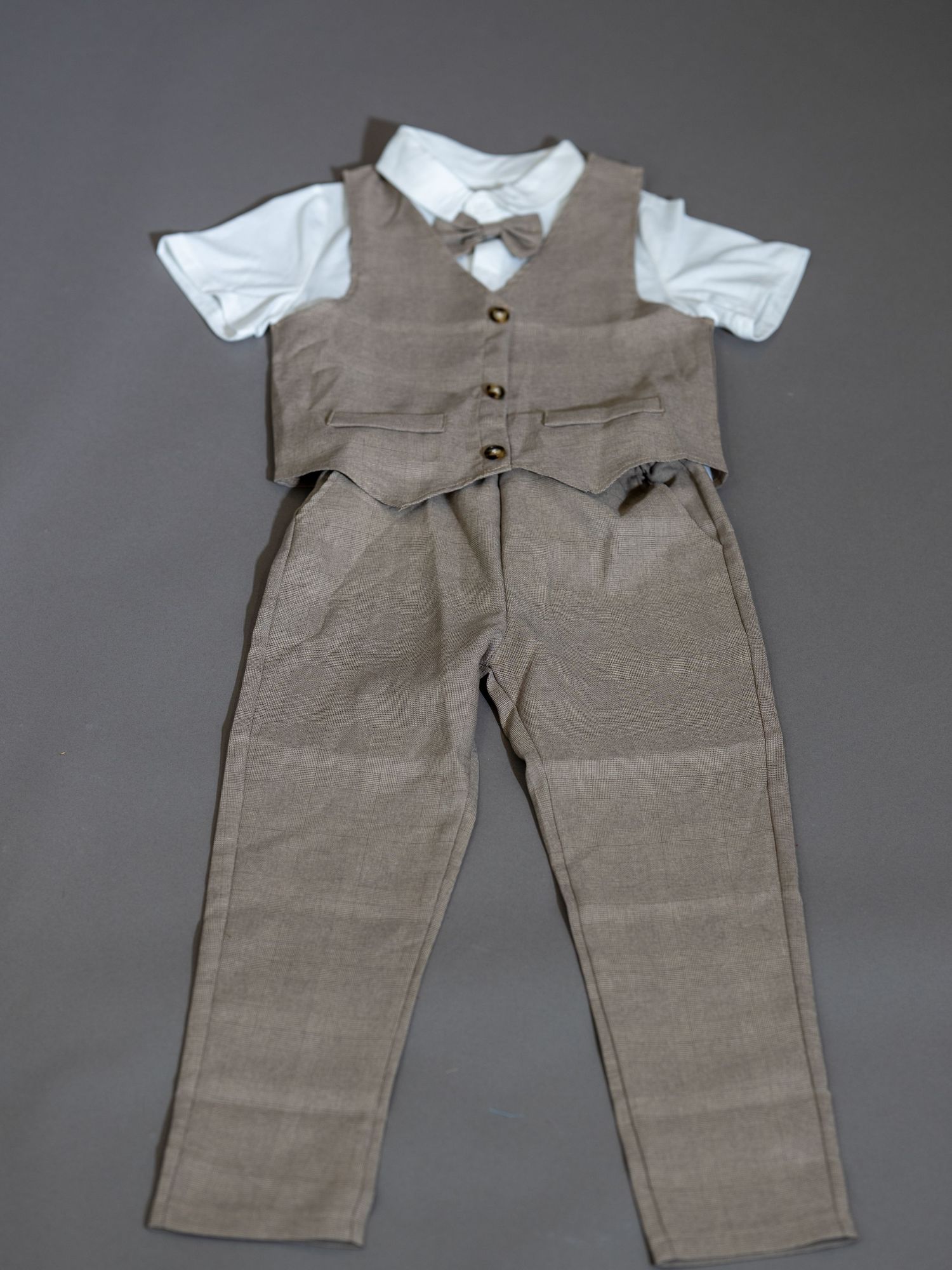 Formell barnoutfit med grått väst och byxor i kombination med vit skjorta och fluga mot mörk bakgrund.