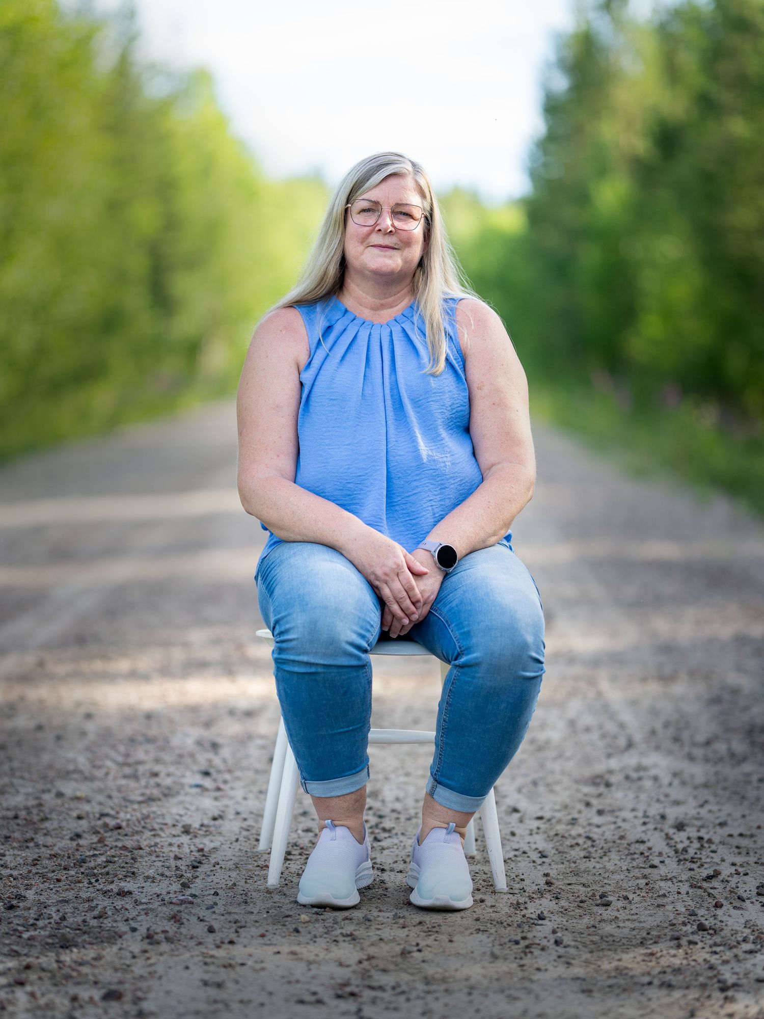 Someone in a blue sleeveless top and jeans sits on a white chair on a dirt path surrounded by green trees.