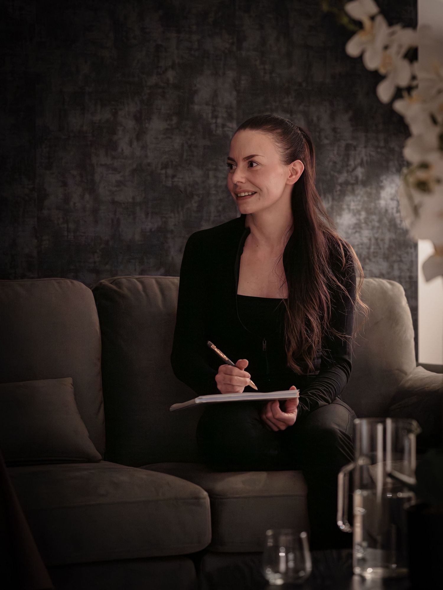 A woman in dark clothing sits on a grey couch writing in a notebook with soft lighting in a moody setting.