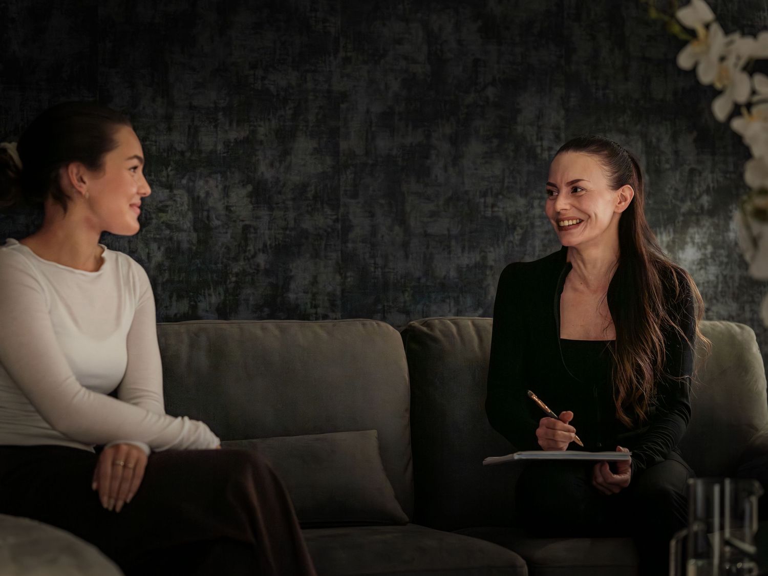 People sit together on a dark gray couch during a relaxed conversation in a dimly lit room.