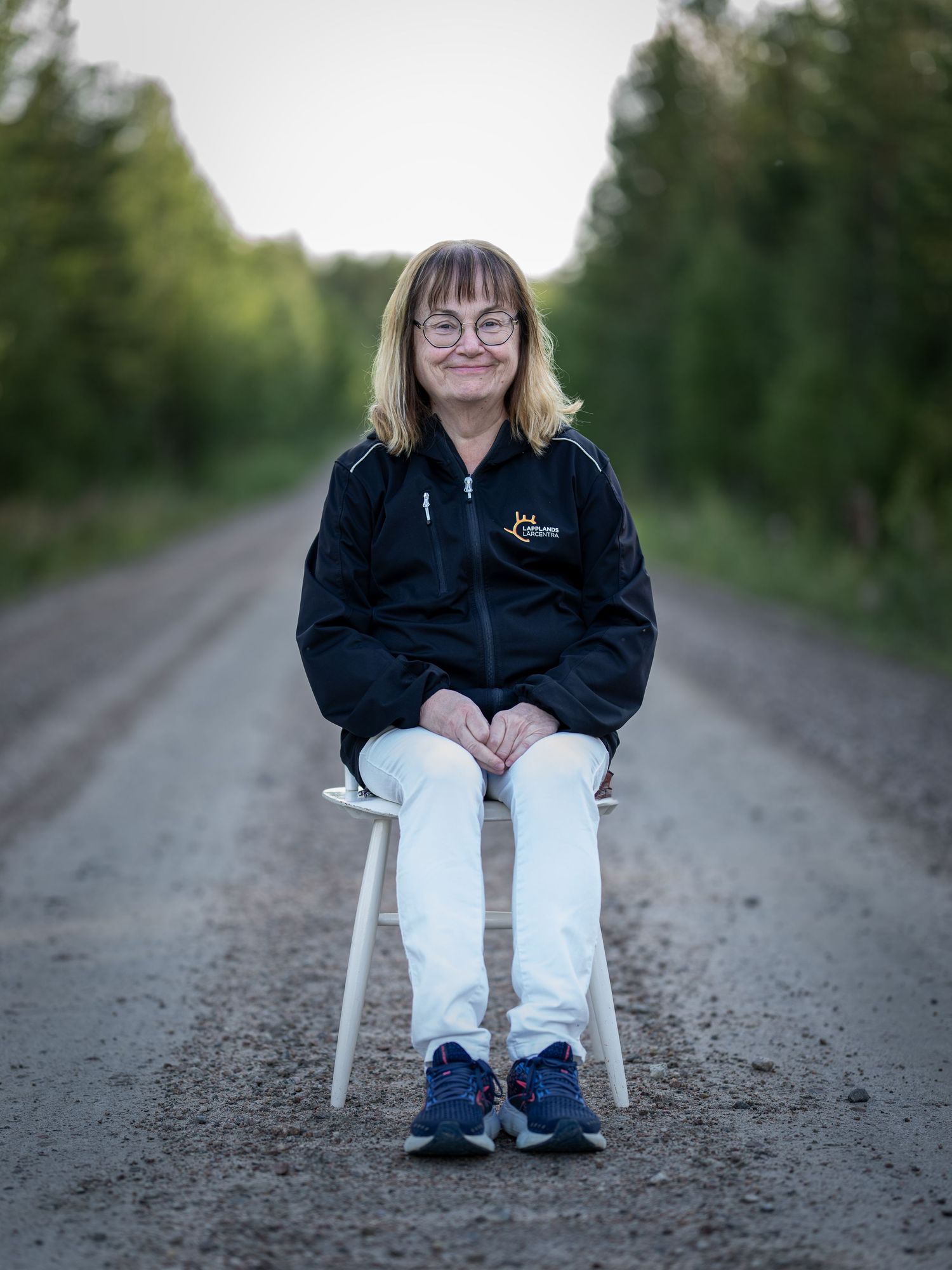 A person wearing a black jacket sits on a white plastic chair on a rural gravel road surrounded by greenery.