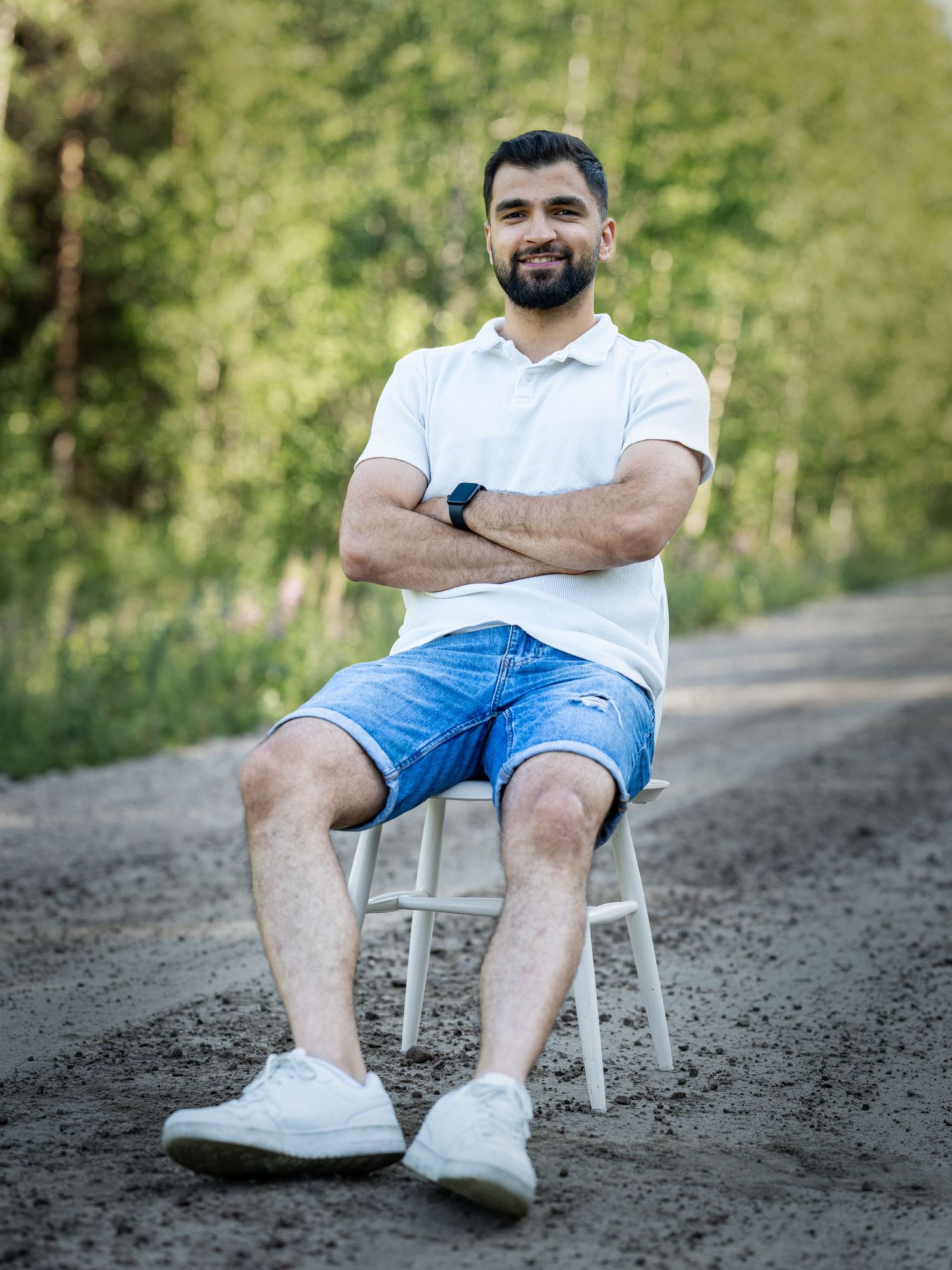 A person in a white shirt and blue shorts sits casually on a white chair on a gravel path surrounded by greenery.