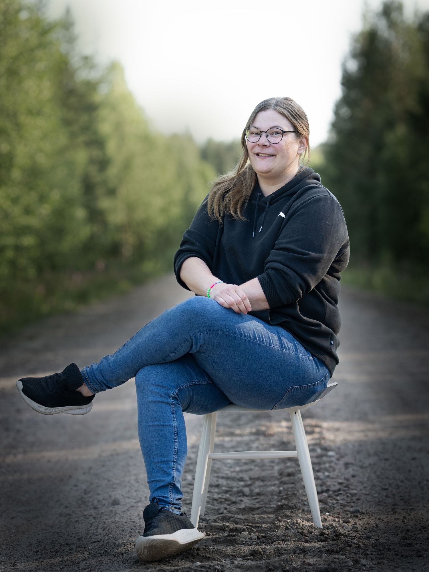A person in jeans and black hoodie sits casually on a wooden stool along a tree-lined path outdoors.